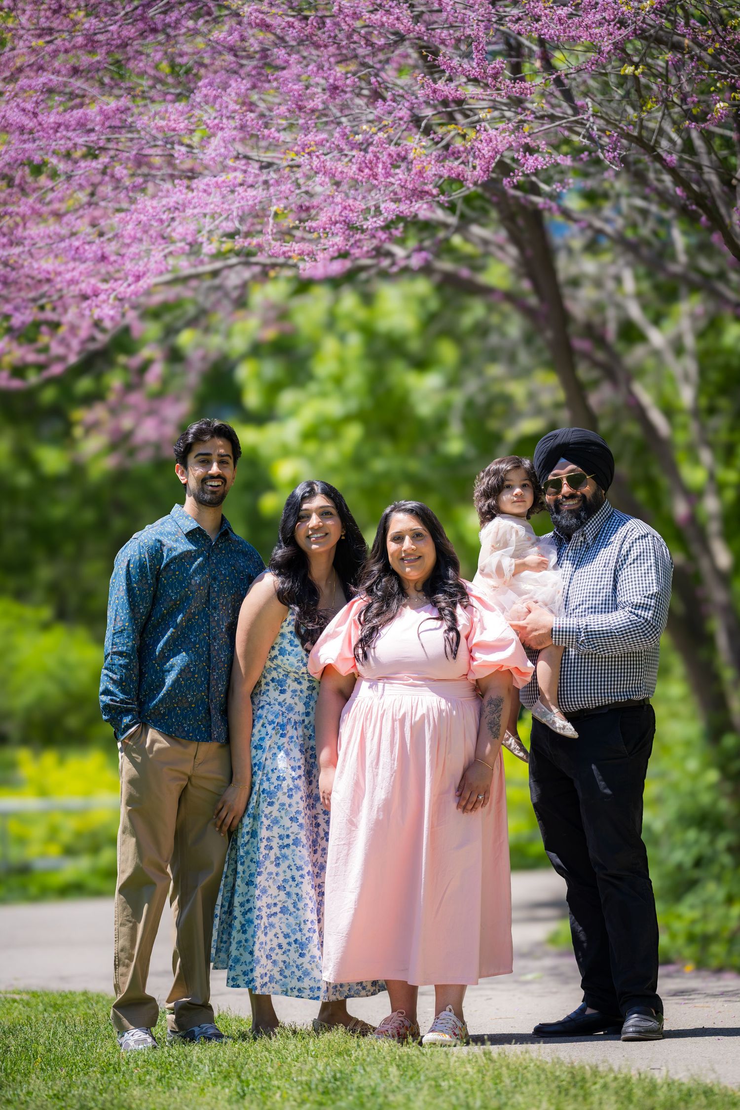 Family of five poses under pink flowering tree in sunny park, mother in pink maternity dress.