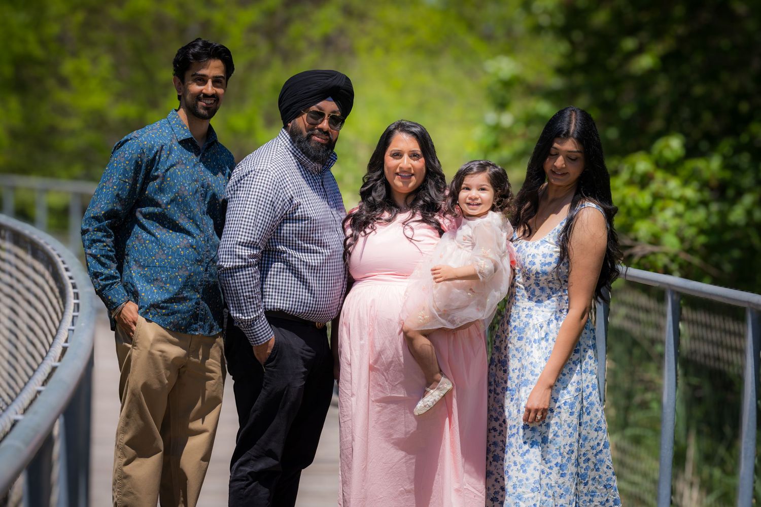 Family of five posing on bridge outdoors, with pregnant woman in pink dress at center.