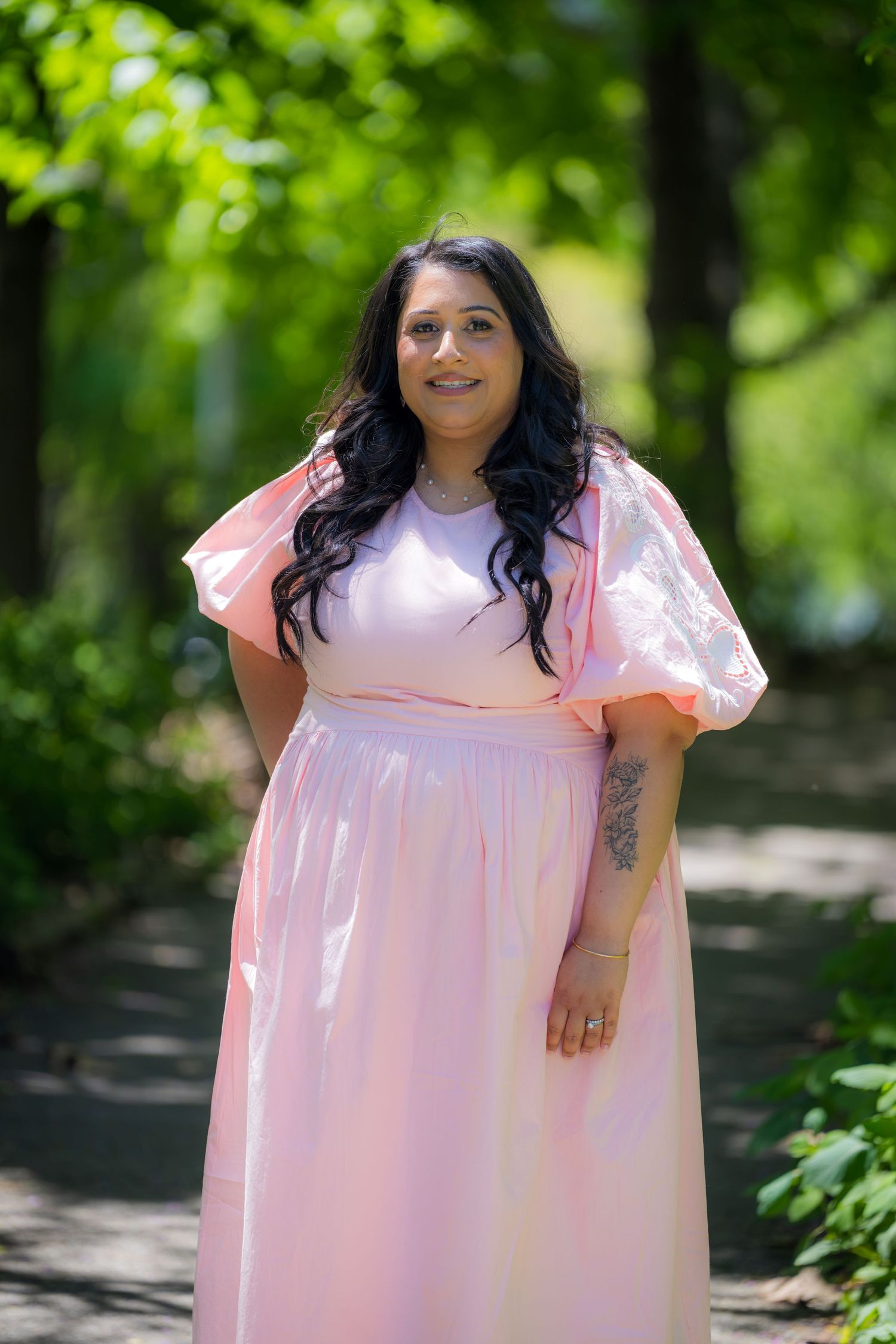Smiling pregnant woman in pink puff sleeve maxi dress stands on garden path among trees.