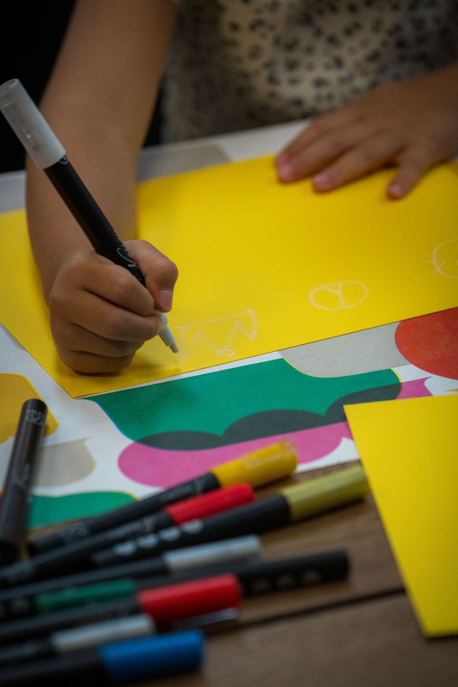 Close-up of child drawing with black marker on yellow paper with colorful shapes and markers nearby.