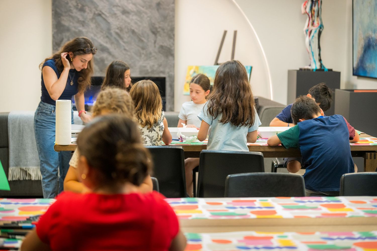 Children engaged in an art class activity at tables in a modern gallery space, supervised by a teacher.