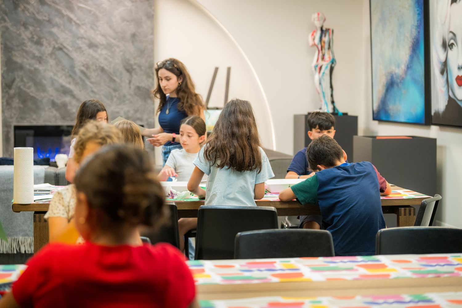 Children engaged in an art class with a teacher guiding students at a table in a modern gallery space.