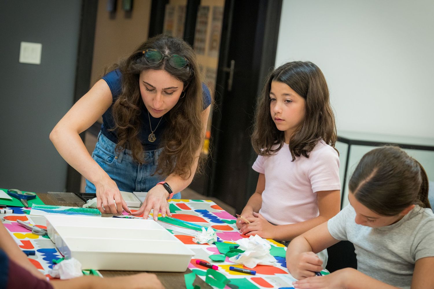 A teacher and two girls focus on a colorful art project at a table.