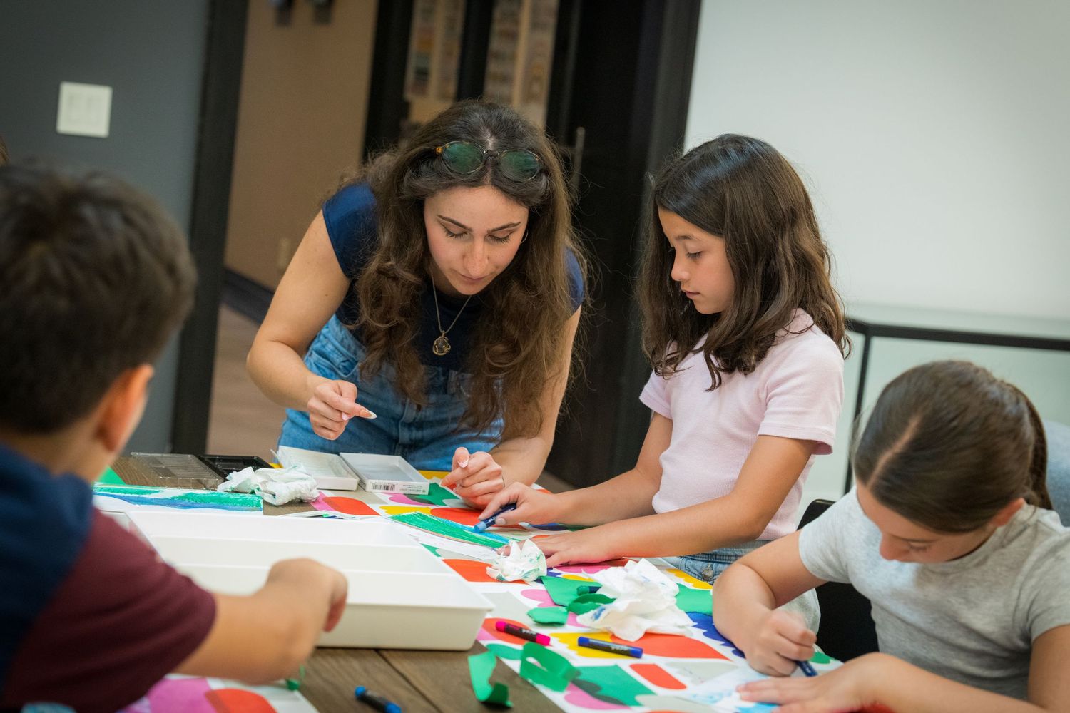 Adult woman guides children in colorful arts and crafts activity at a table covered with bright paper shapes.