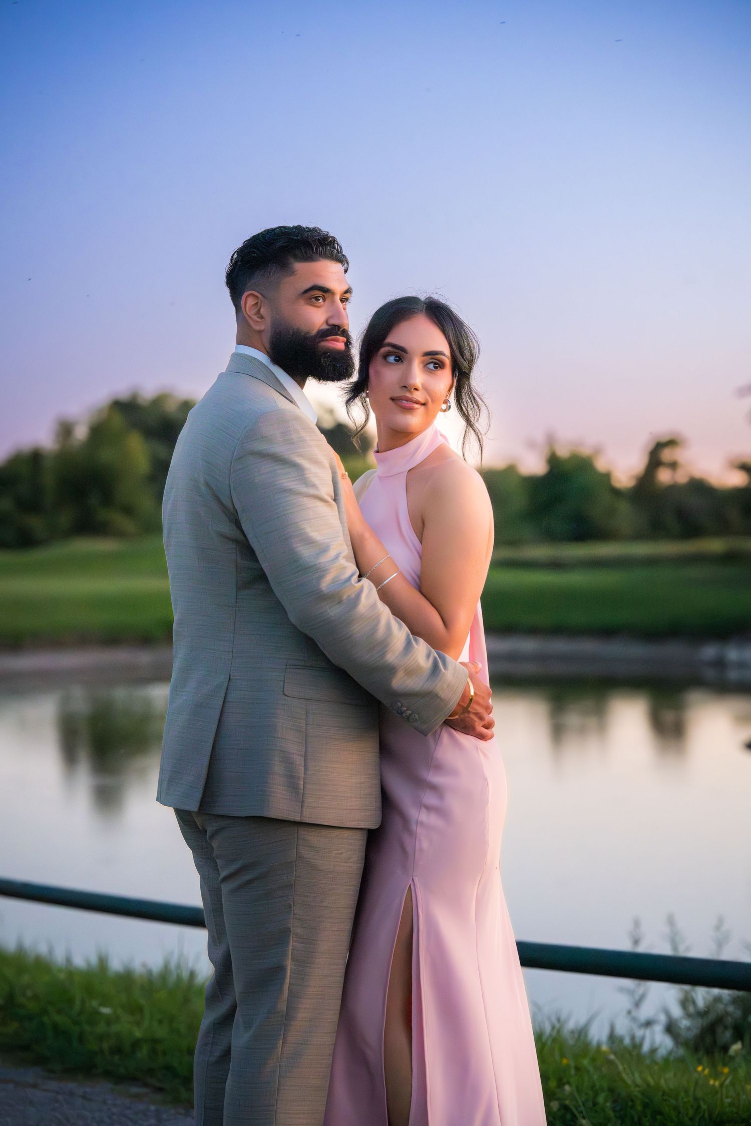 Couple in formal attire embraces during lakeside sunset photoshoot.