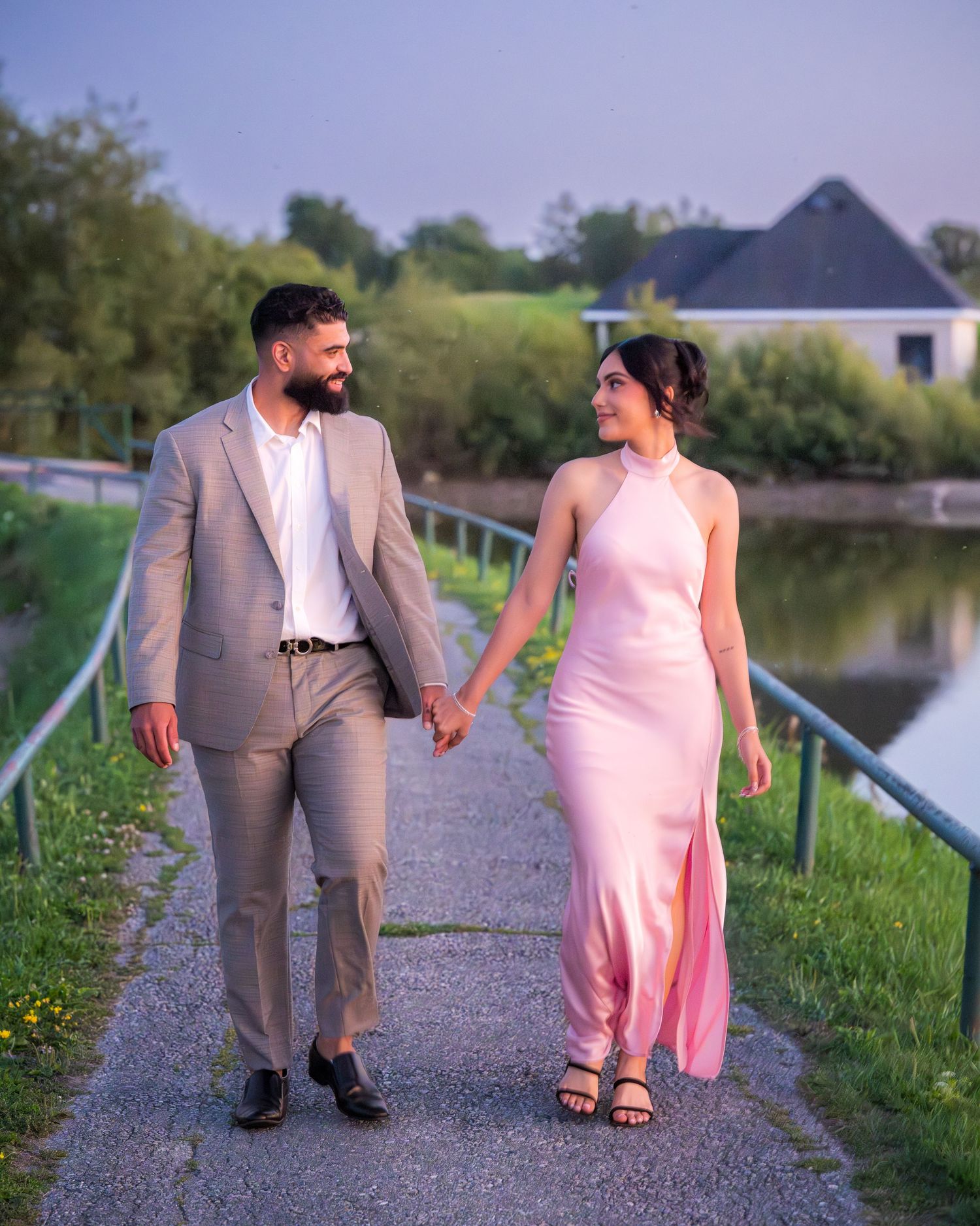 A couple walks together on a waterside path during golden hour, dressed in formal attire.