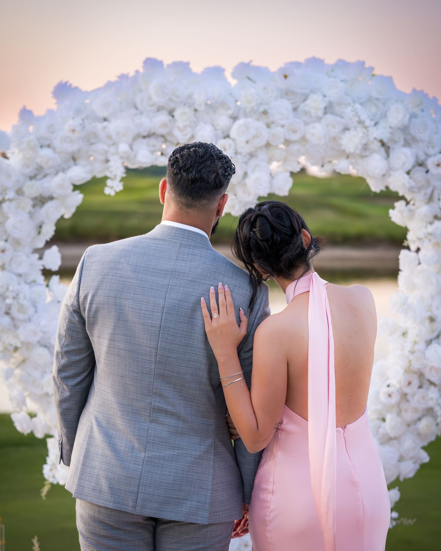 A couple stands before a white floral arch during a romantic outdoor photoshoot.