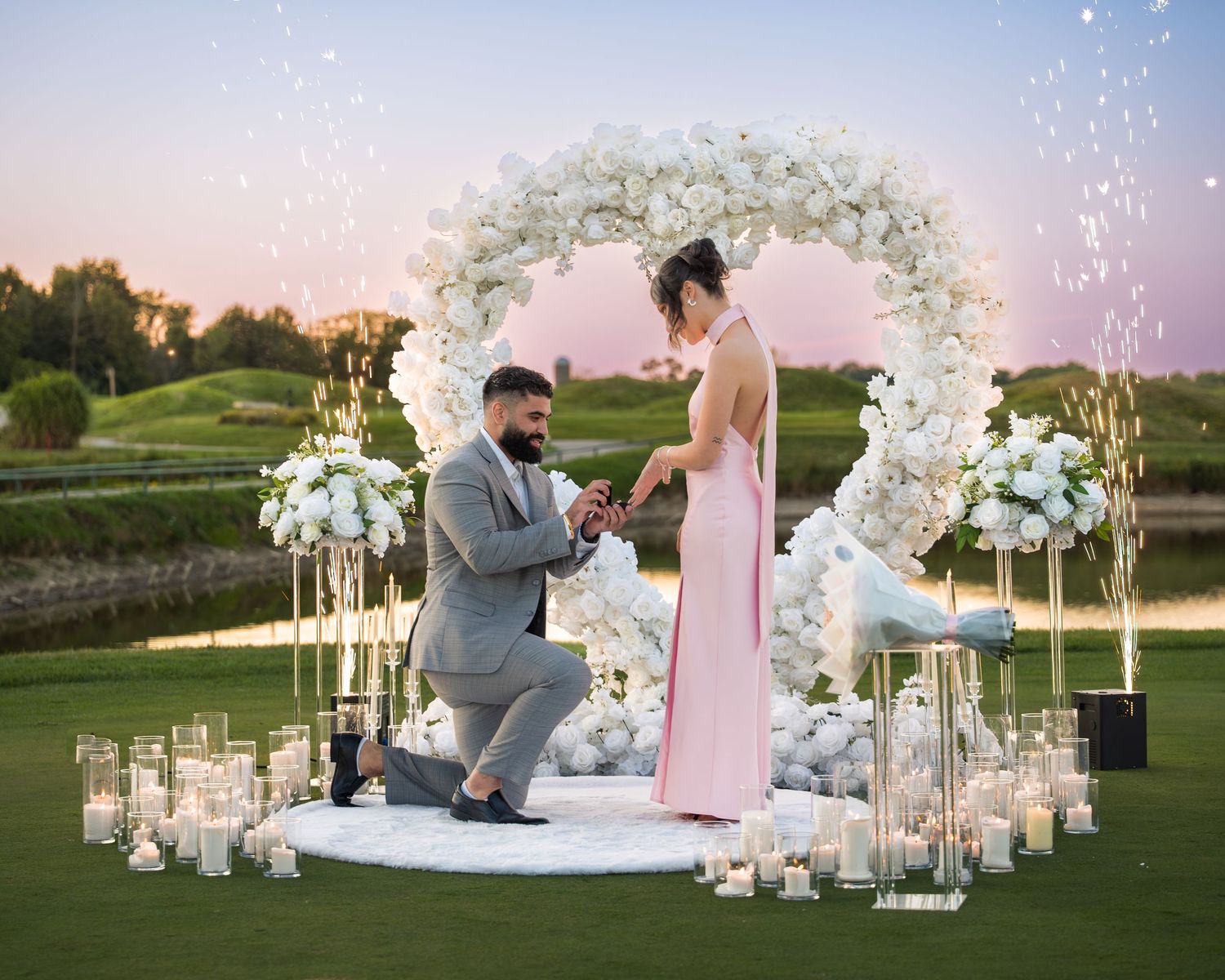 Romantic proposal scene with white orchid arch and candlelit path at sunset.