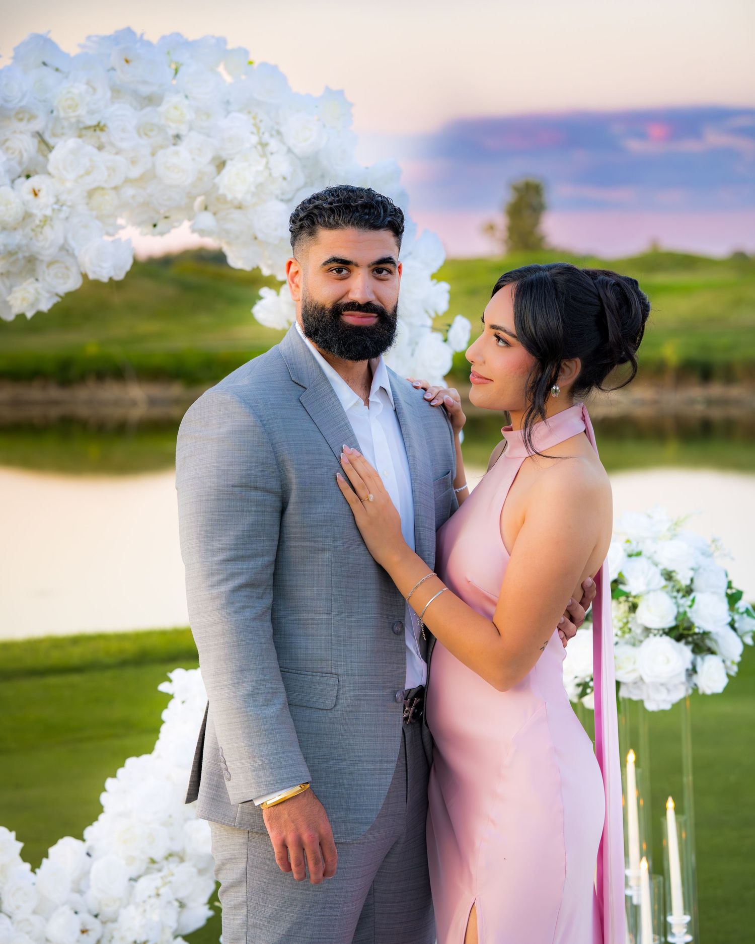 A romantic outdoor portrait of a couple beneath a white floral arch during golden hour.