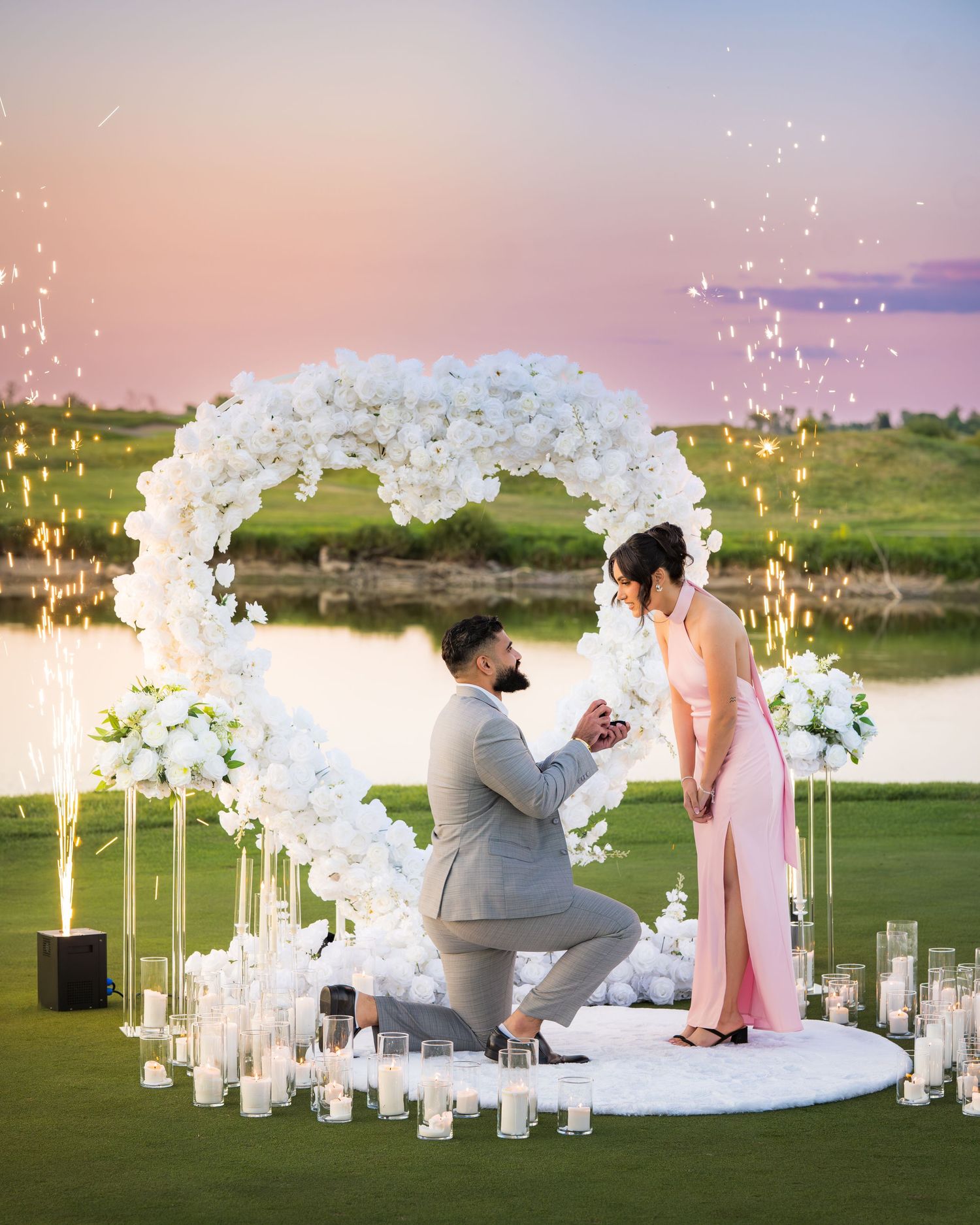 Romantic proposal scene with white flower arch and glowing candles on grass.