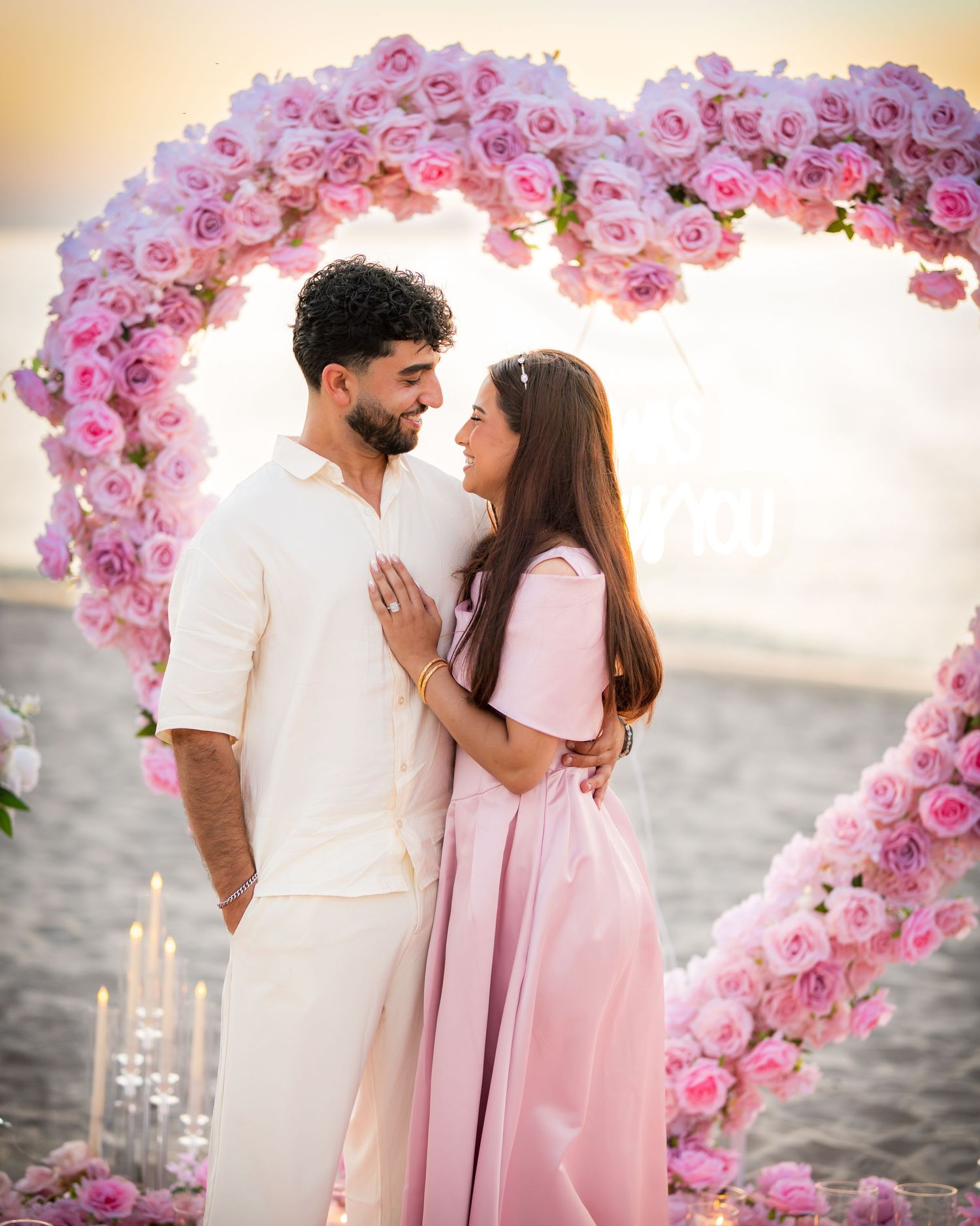 Romantic beach proposal setup with heart-shaped pink rose arch and candles at sunset.