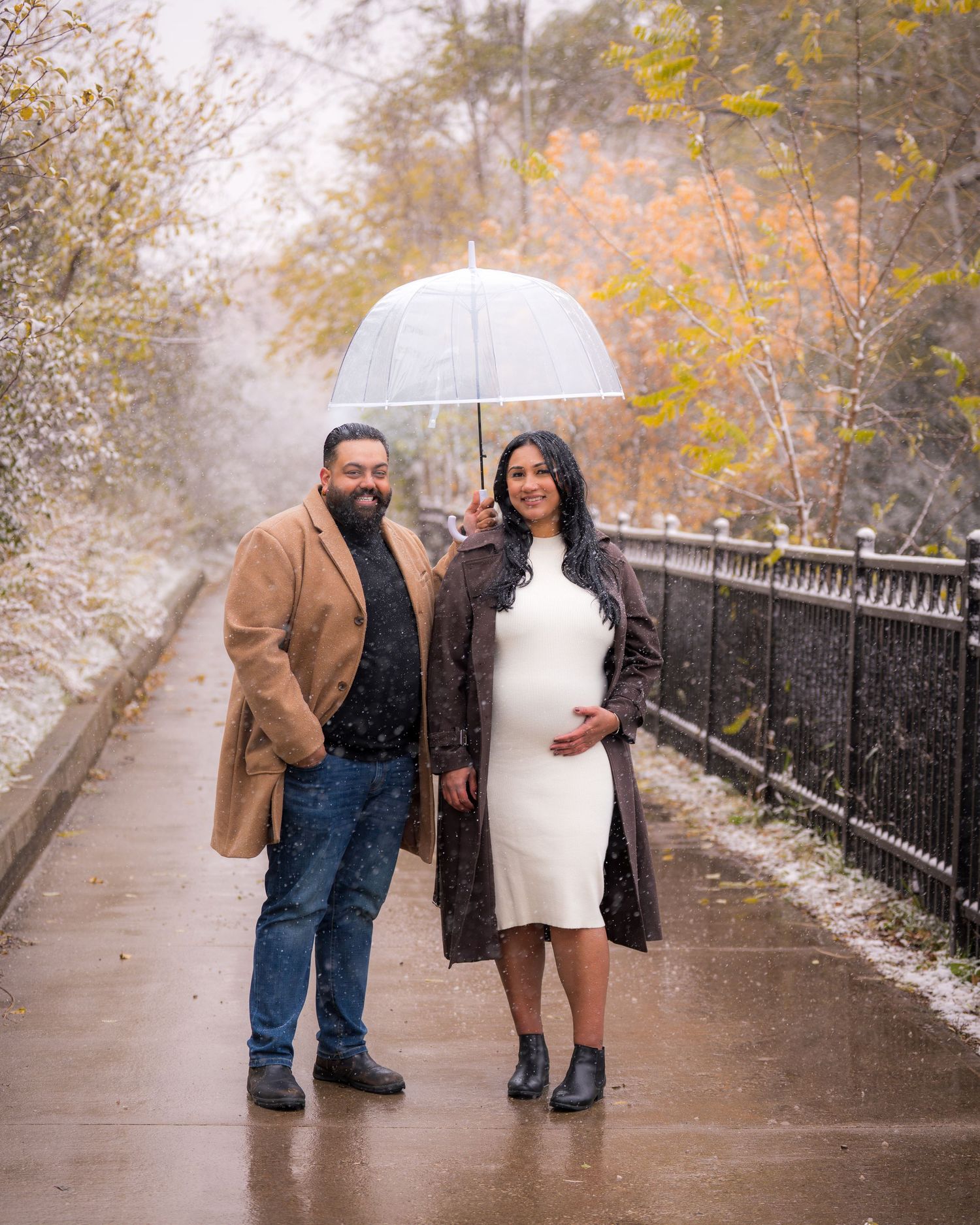 A couple poses on a pathway surrounded by fall foliage and light snow.