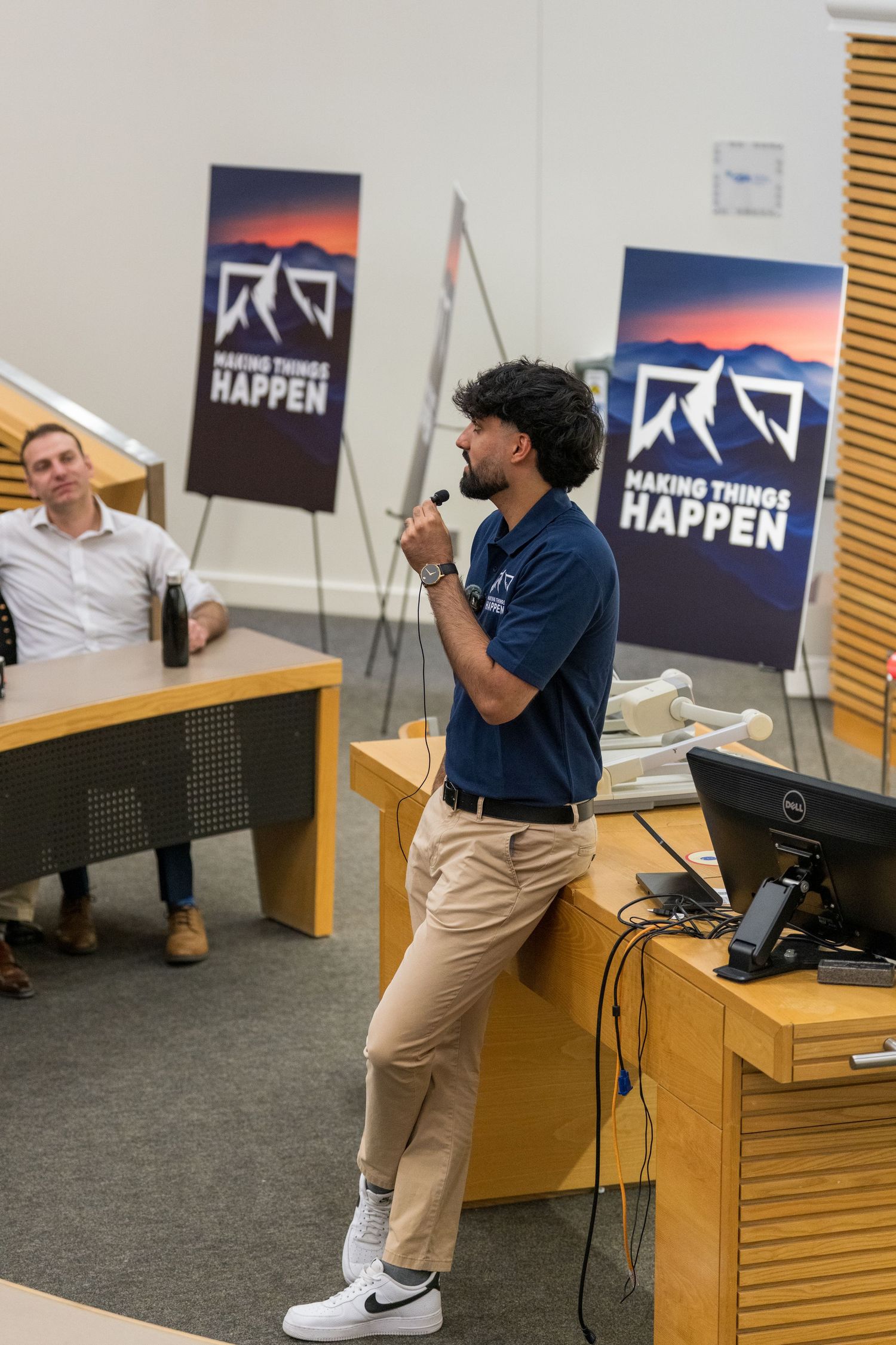 A speaker in a navy polo and khaki pants presents in front of a podium in a conference room.