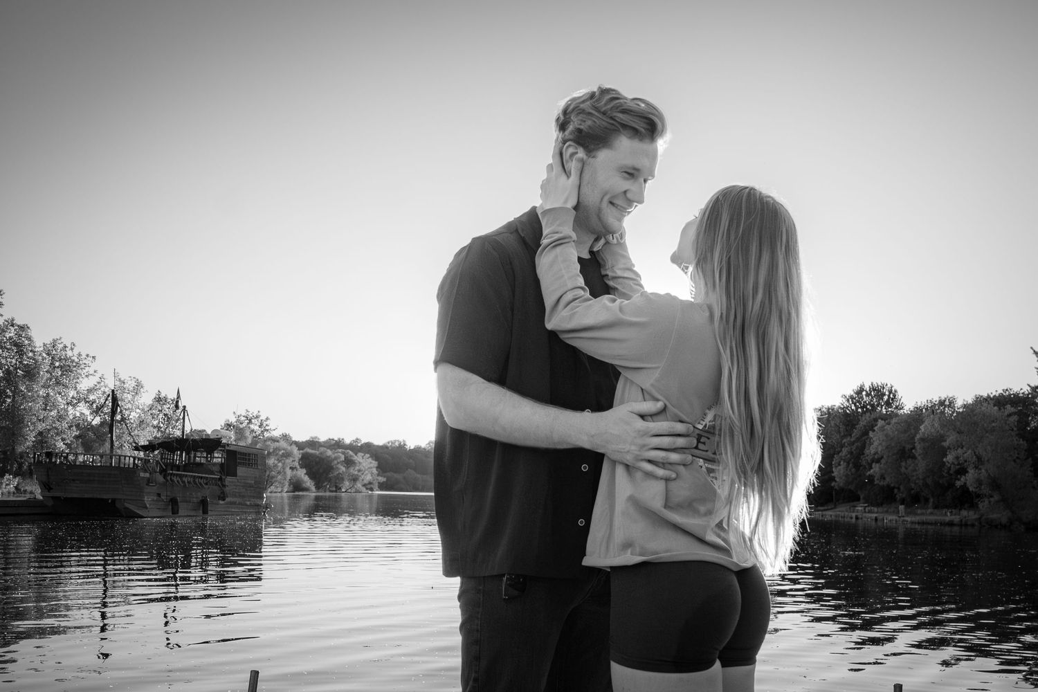 Black and white image of smiling couple hugging closely near a calm lakeside.