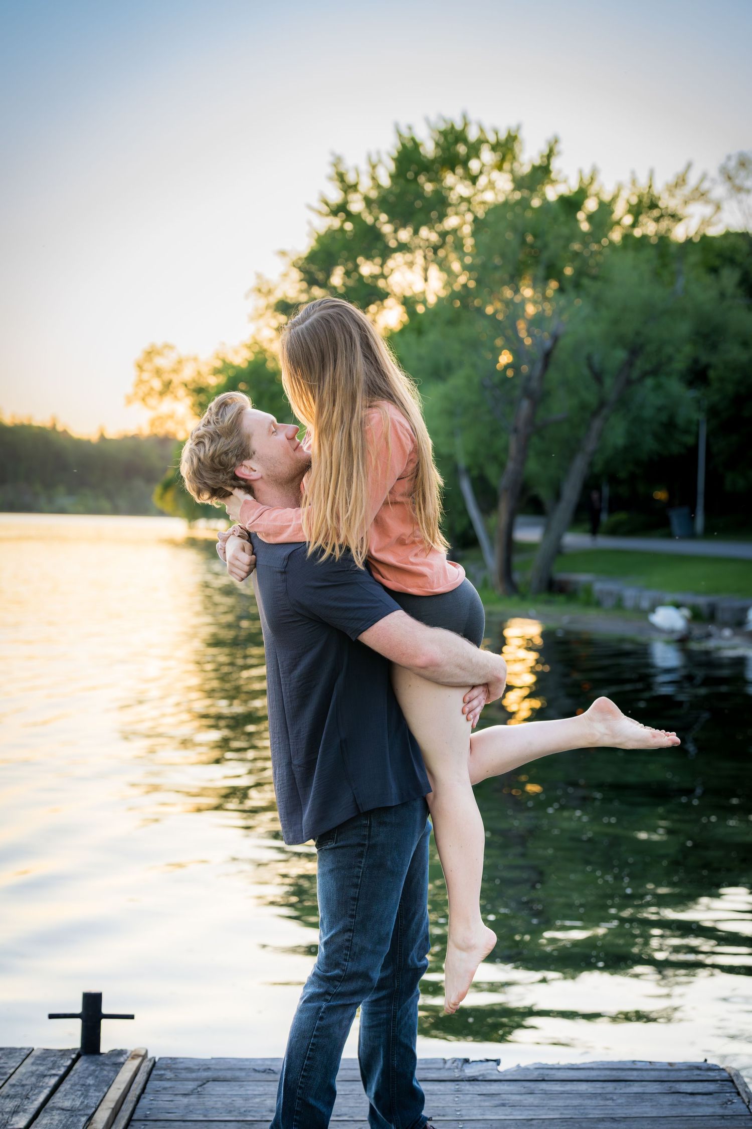 Couple sharing romantic moment at sunset by lake, man lifting woman on wooden dock.