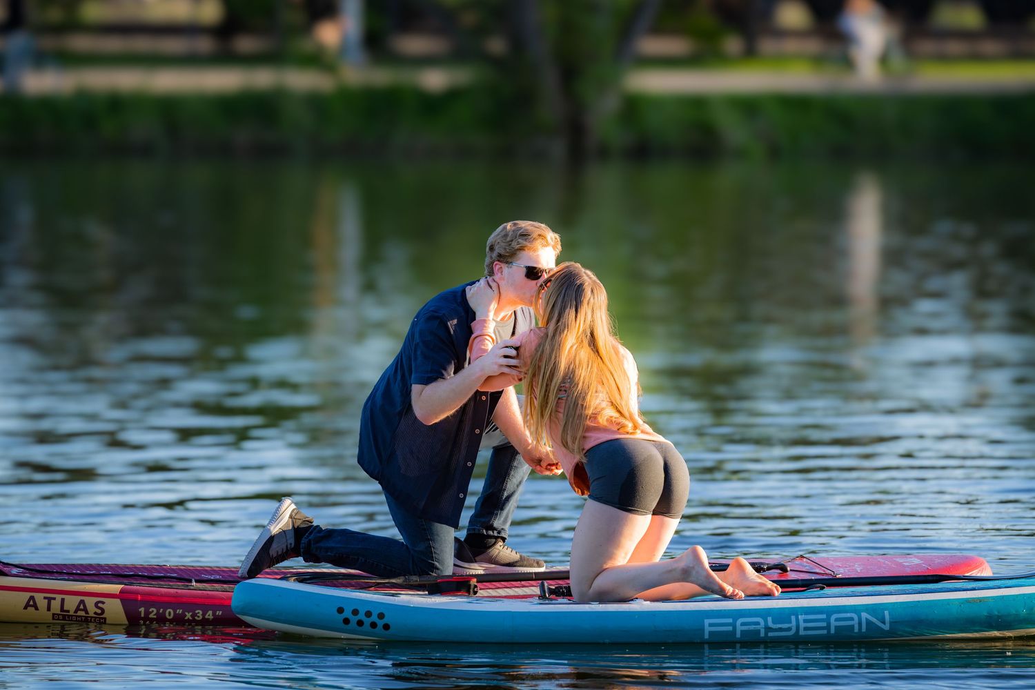 Couple sharing a kiss on a paddleboard on a calm lake during a romantic proposal moment.