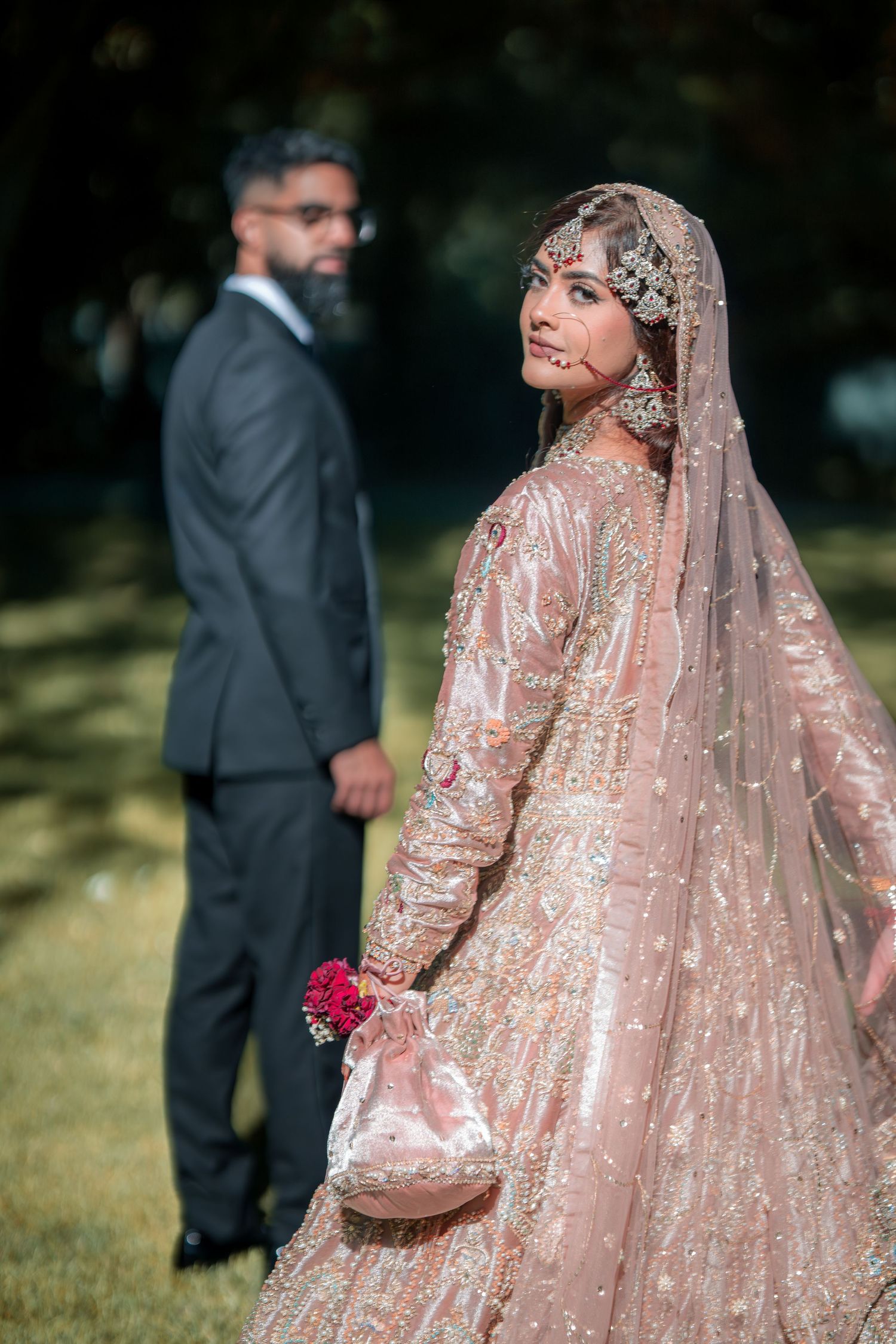 Bride in glittering rose gold lehenga poses gracefully in an outdoor garden setting.