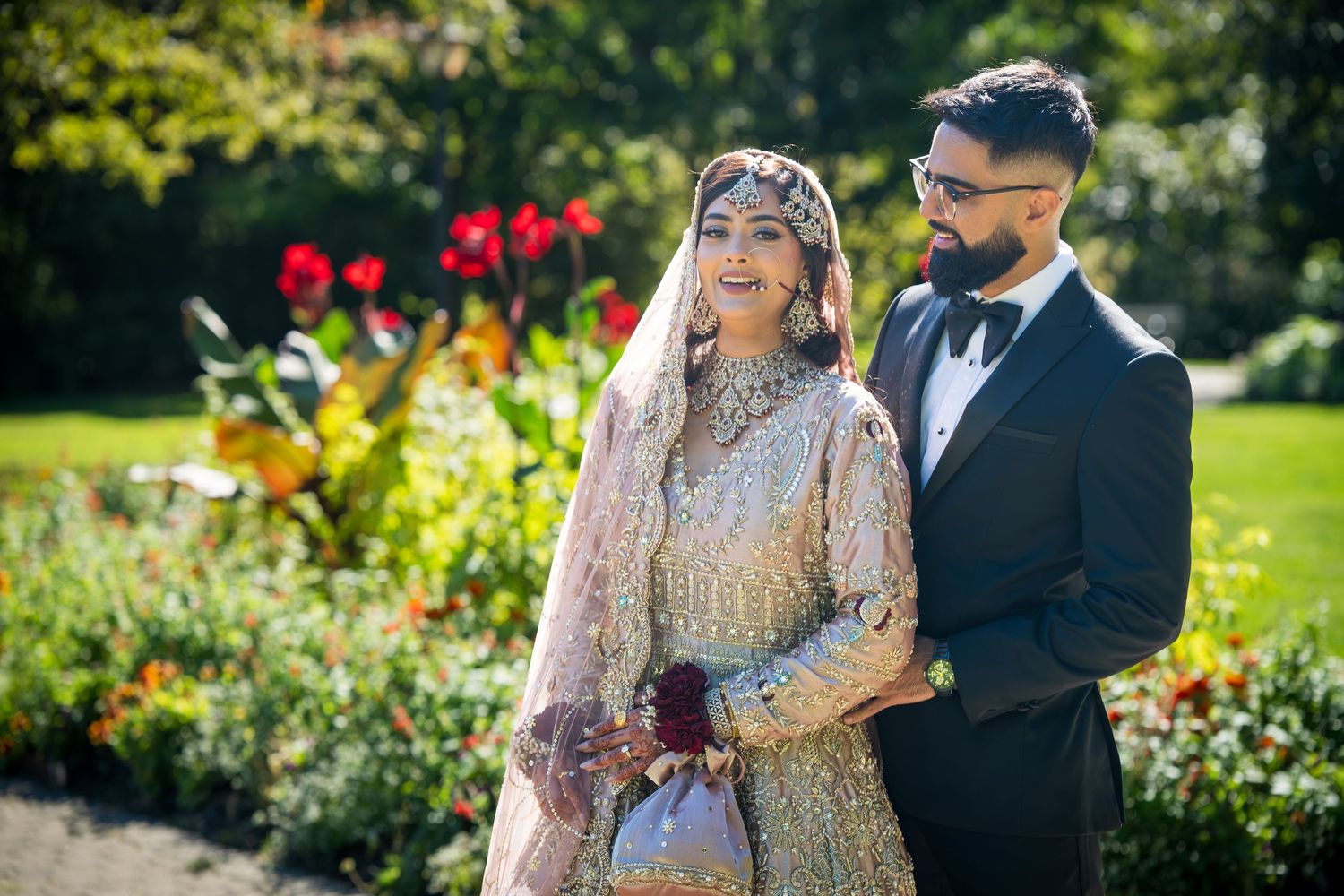 A couple in traditional South Asian wedding attire pose in a garden with red flowers in the background.