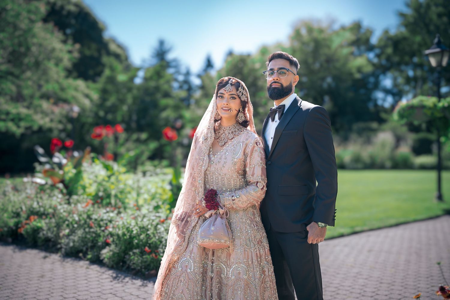 A wedding couple poses outdoors with the bride in an embellished lehenga and groom in a classic tuxedo.