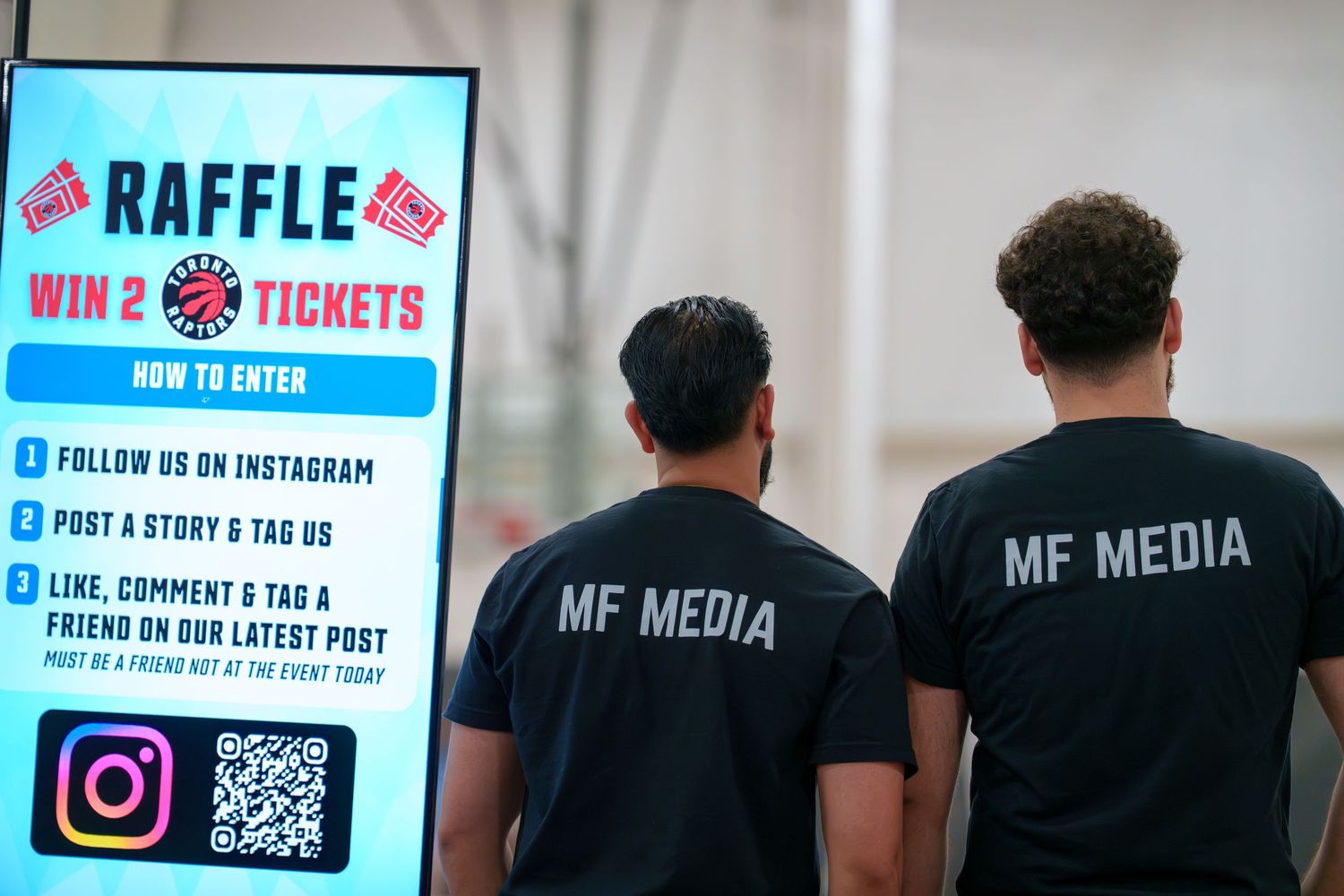 Two people wearing black MF Media t-shirts stand in front of a blue raffle sign with social media instructions.