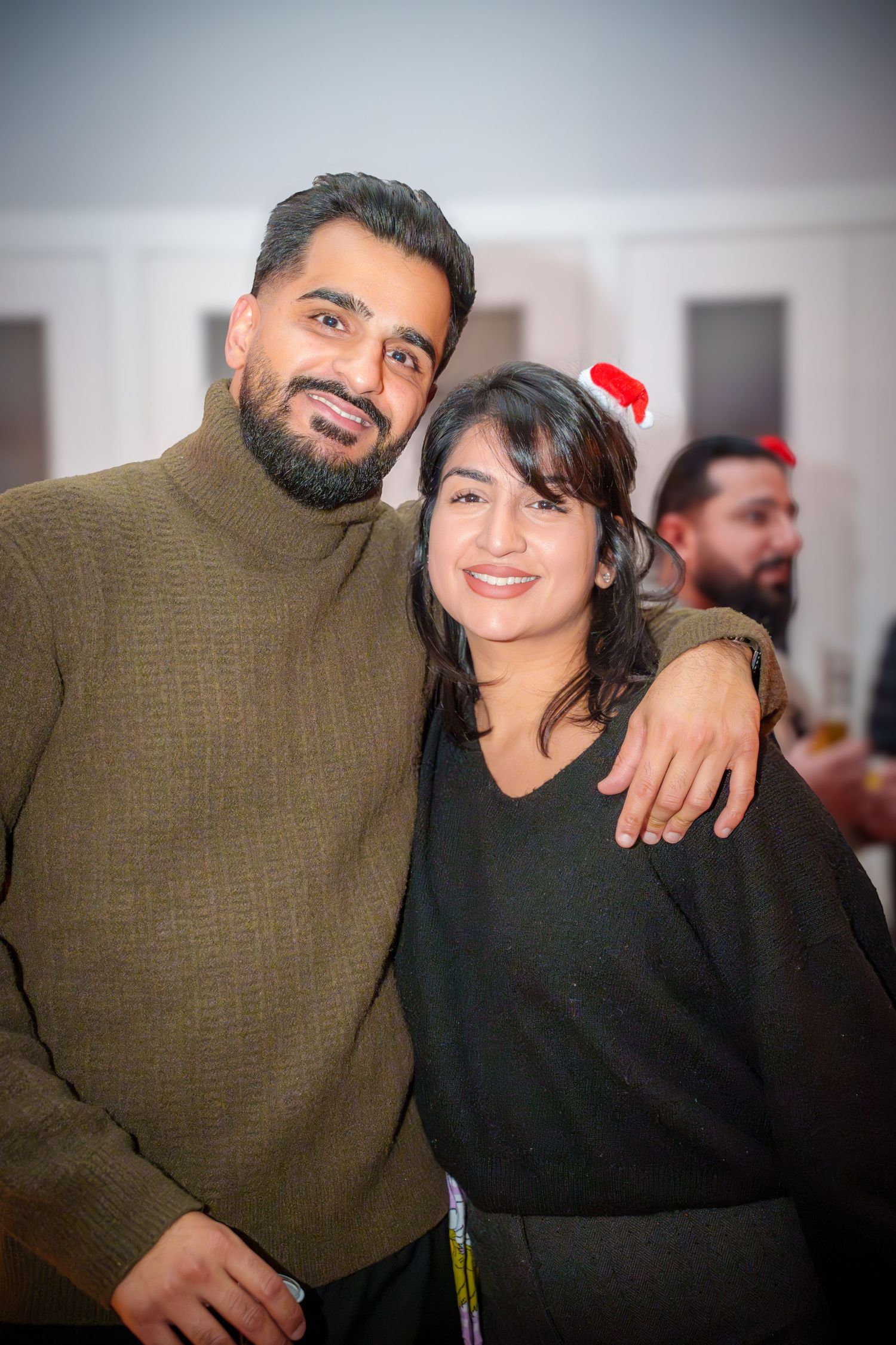 Couple in casual attire smiling at holiday party with festive decorations visible in background.