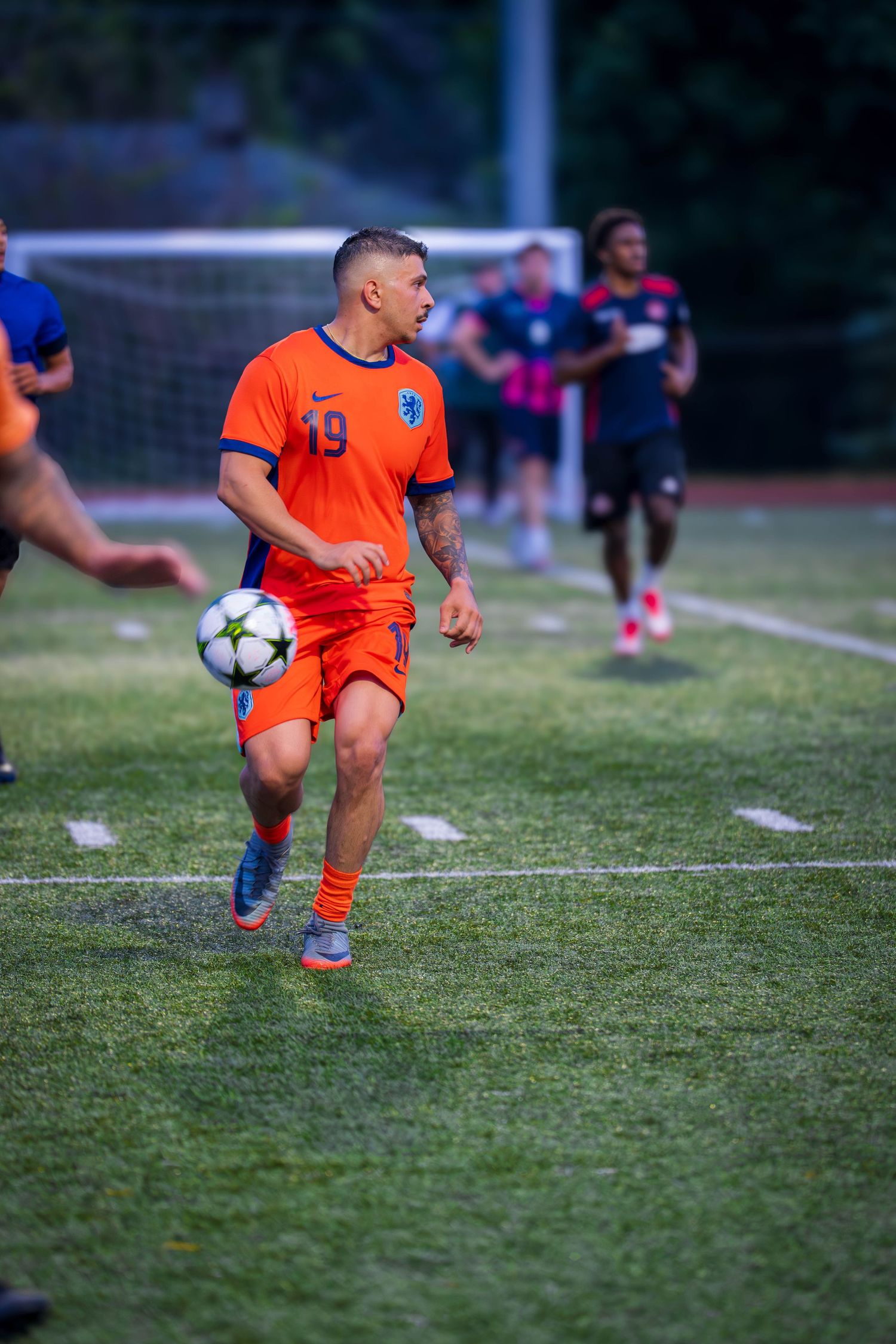 Player in orange uniform controls soccer ball during game with goal visible in background.