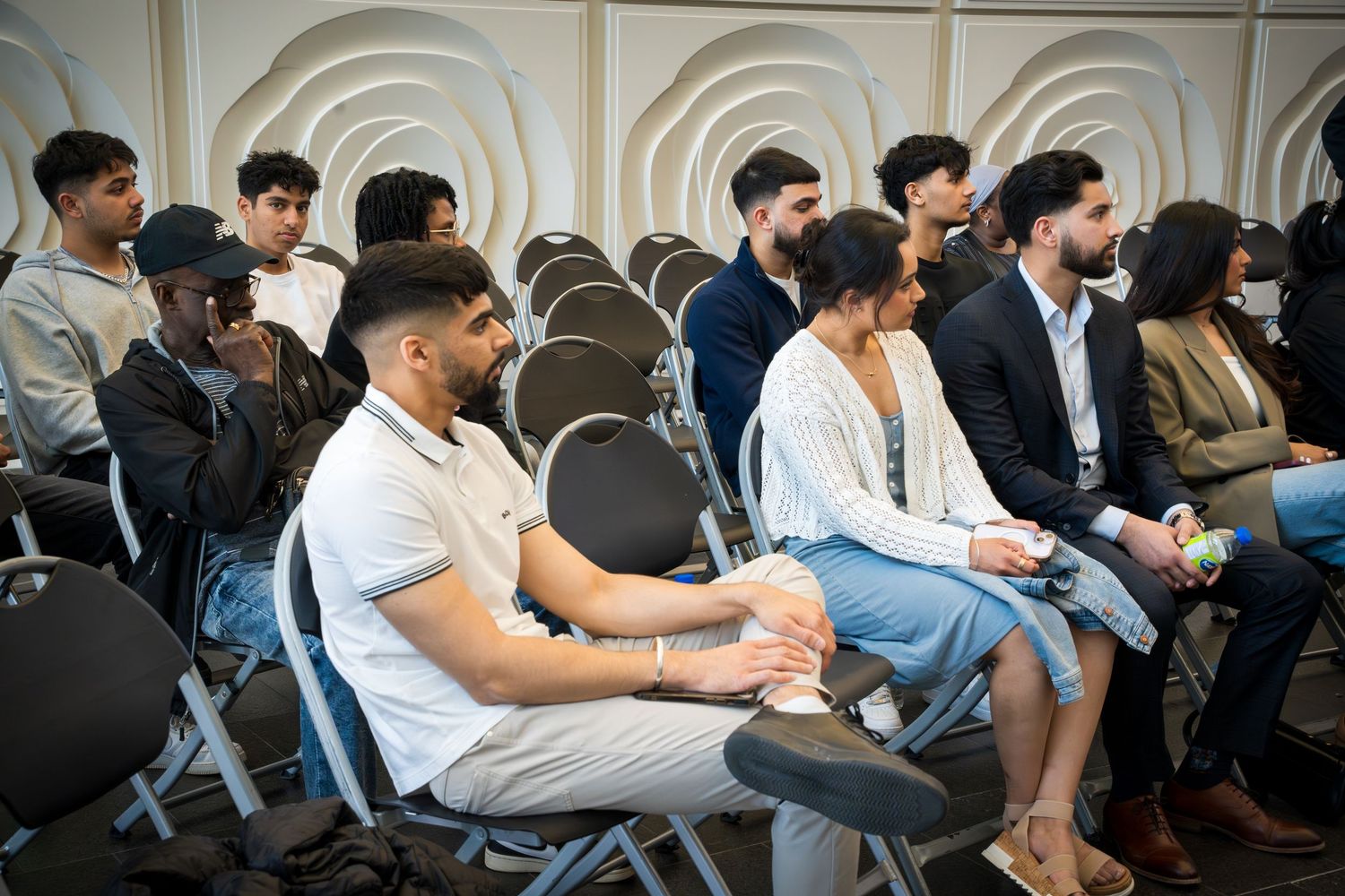 Attendees seated in rows of folding chairs against white textured wall during indoor event.