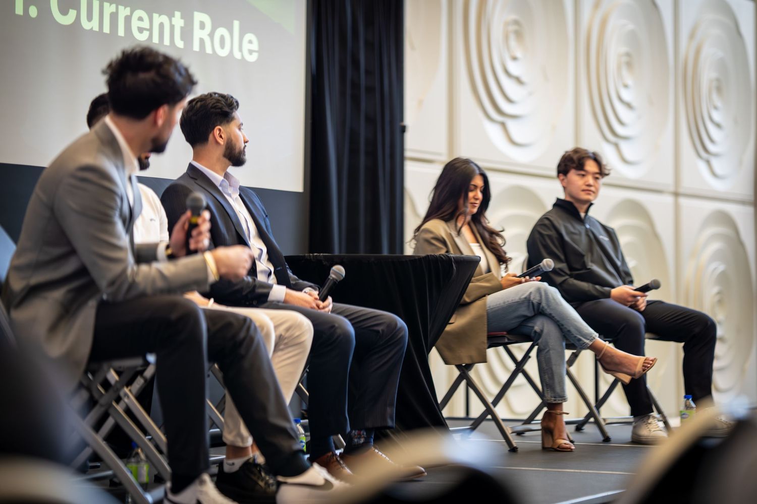 Panel discussion participants seated in folding chairs on stage with black curtain backdrop and white textured wall.