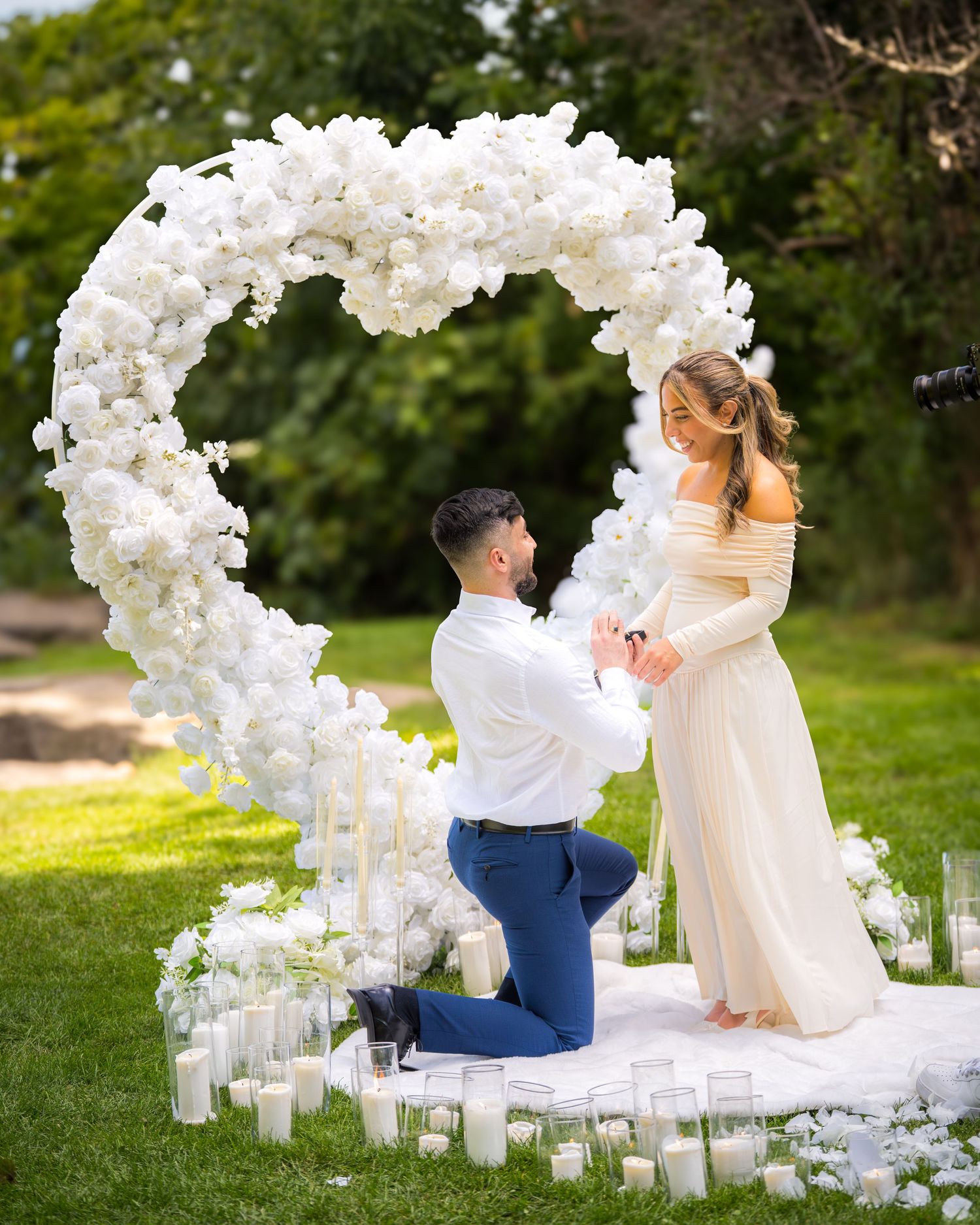 A romantic garden proposal setup with white floral arches and scattered candles on the ground.