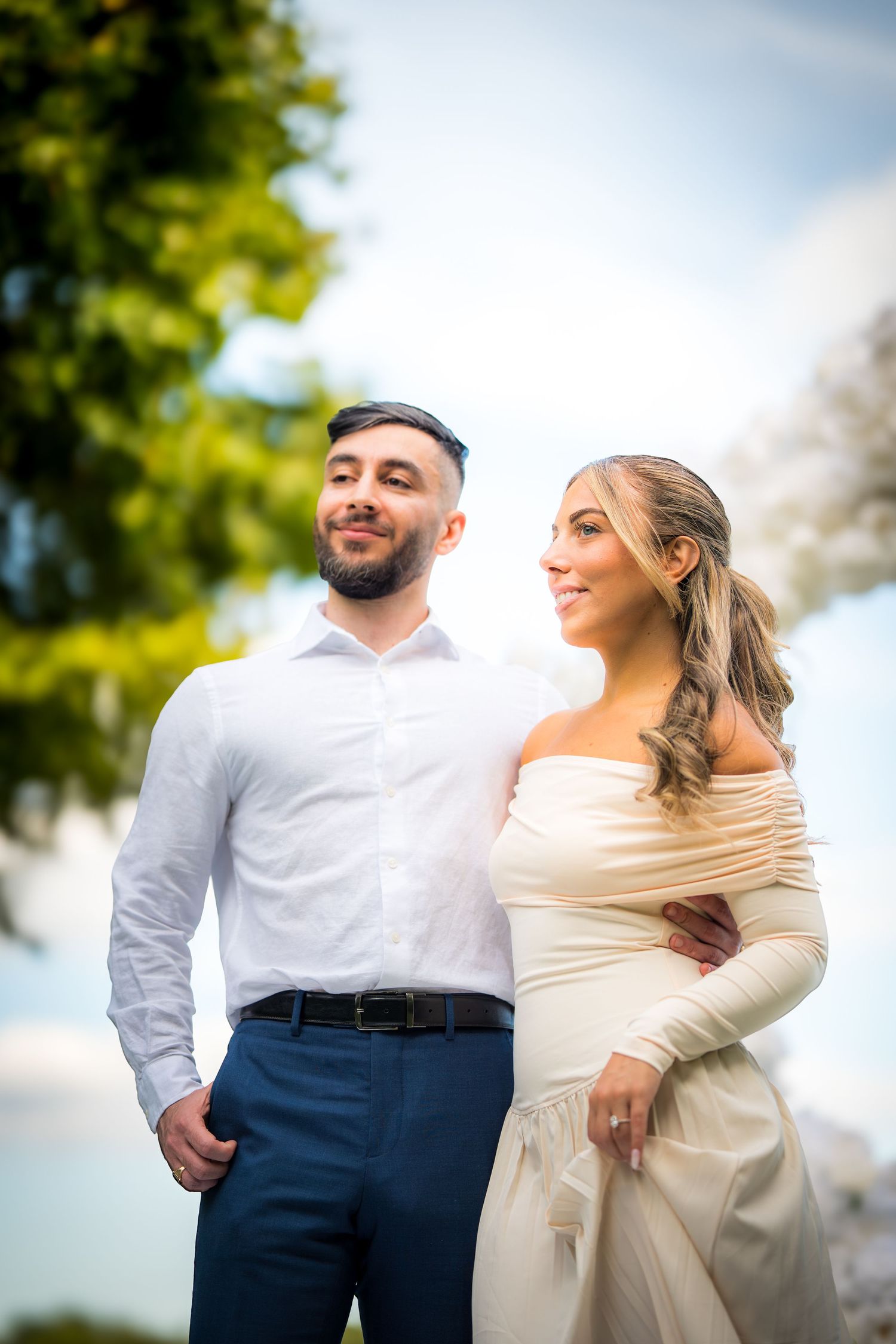 A couple poses together outdoors in formal attire against a sunny natural background.