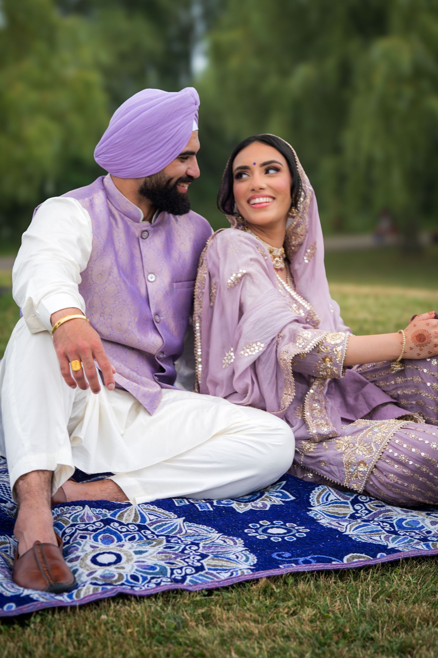 A couple in coordinating purple and white traditional Indian outfits relax on a blue patterned blanket outdoors.