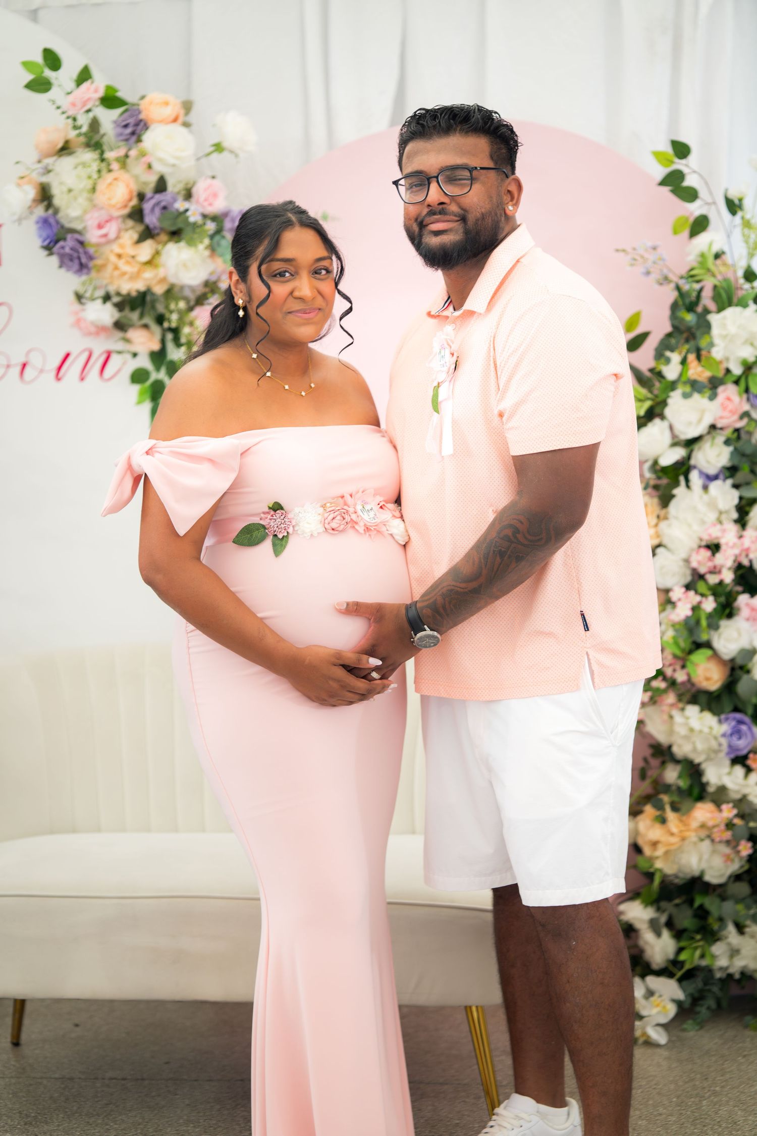 A couple stands together in coordinated outfits against a backdrop of roses and greenery.