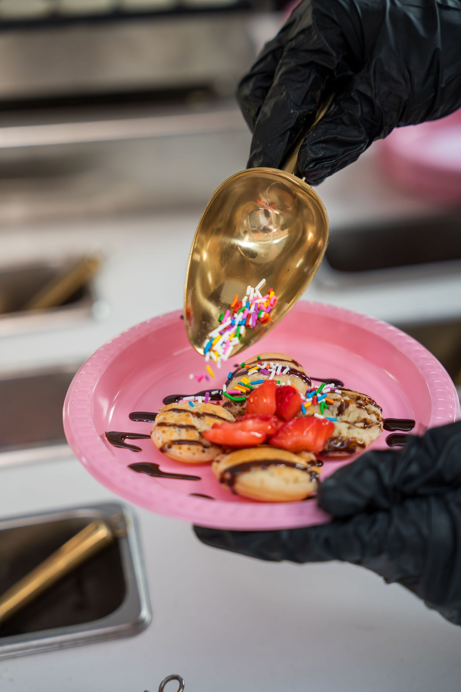 Chef in black gloves plating desserts on pink plates with colorful toppings and garnishes.