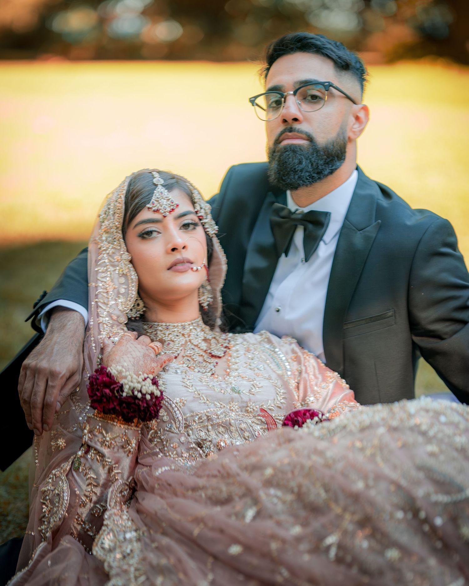 A romantic wedding portrait of a couple in traditional South Asian attire against a warm sunset background.