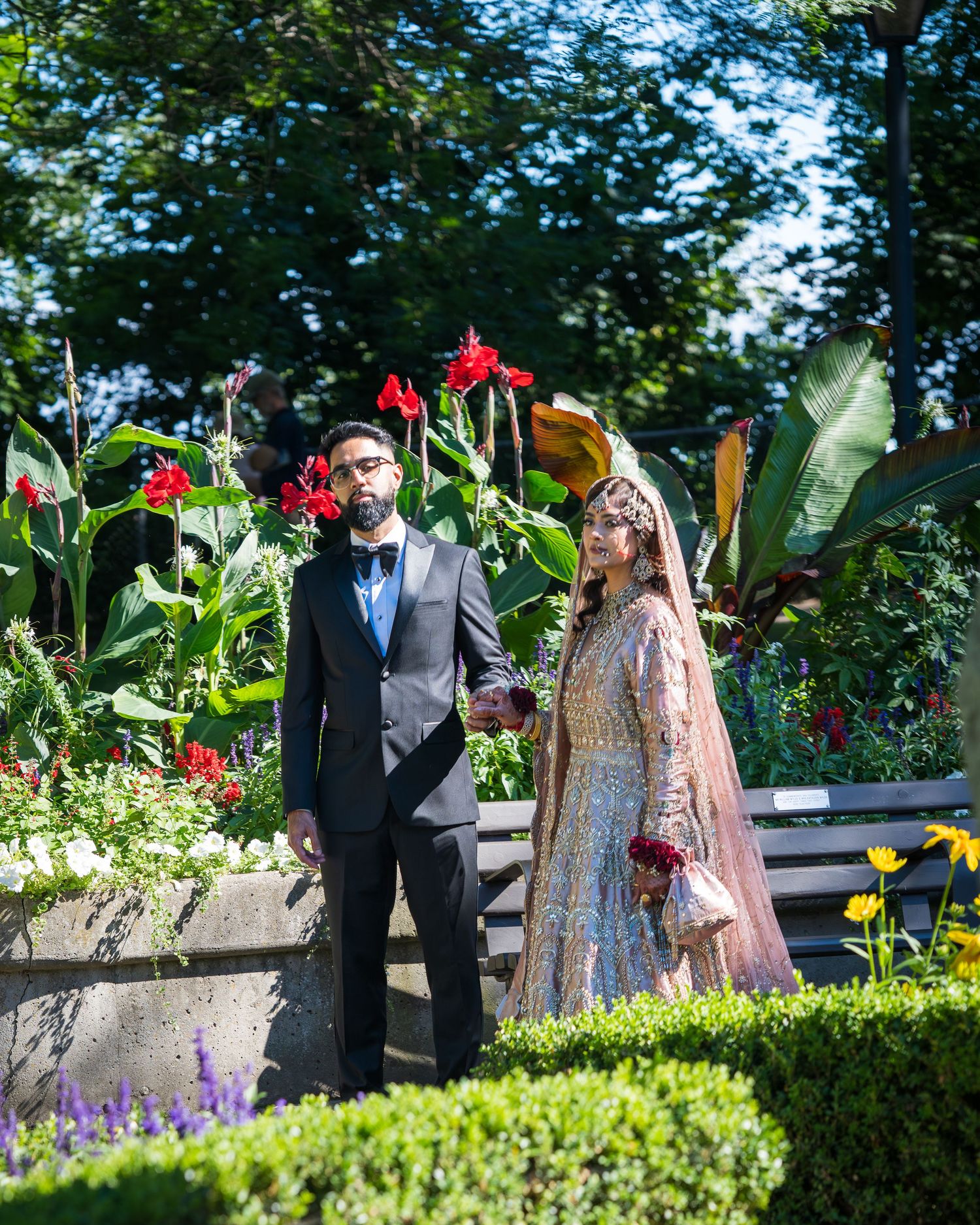 Wedding couple takes romantic portraits in a manicured garden with red flowers and hedges.