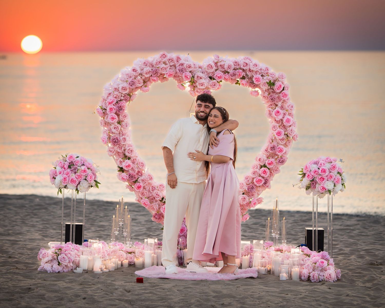 A beach wedding ceremony setup with pink floral arches and decorations against an orange sunset over the ocean.