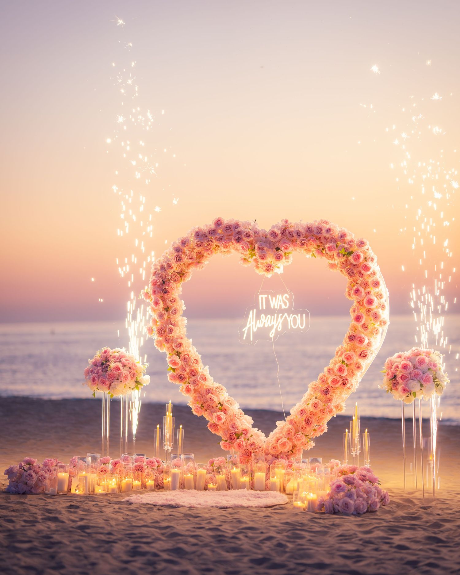 A heart-shaped floral arch with sparklers and candles glowing on a beach at dusk.