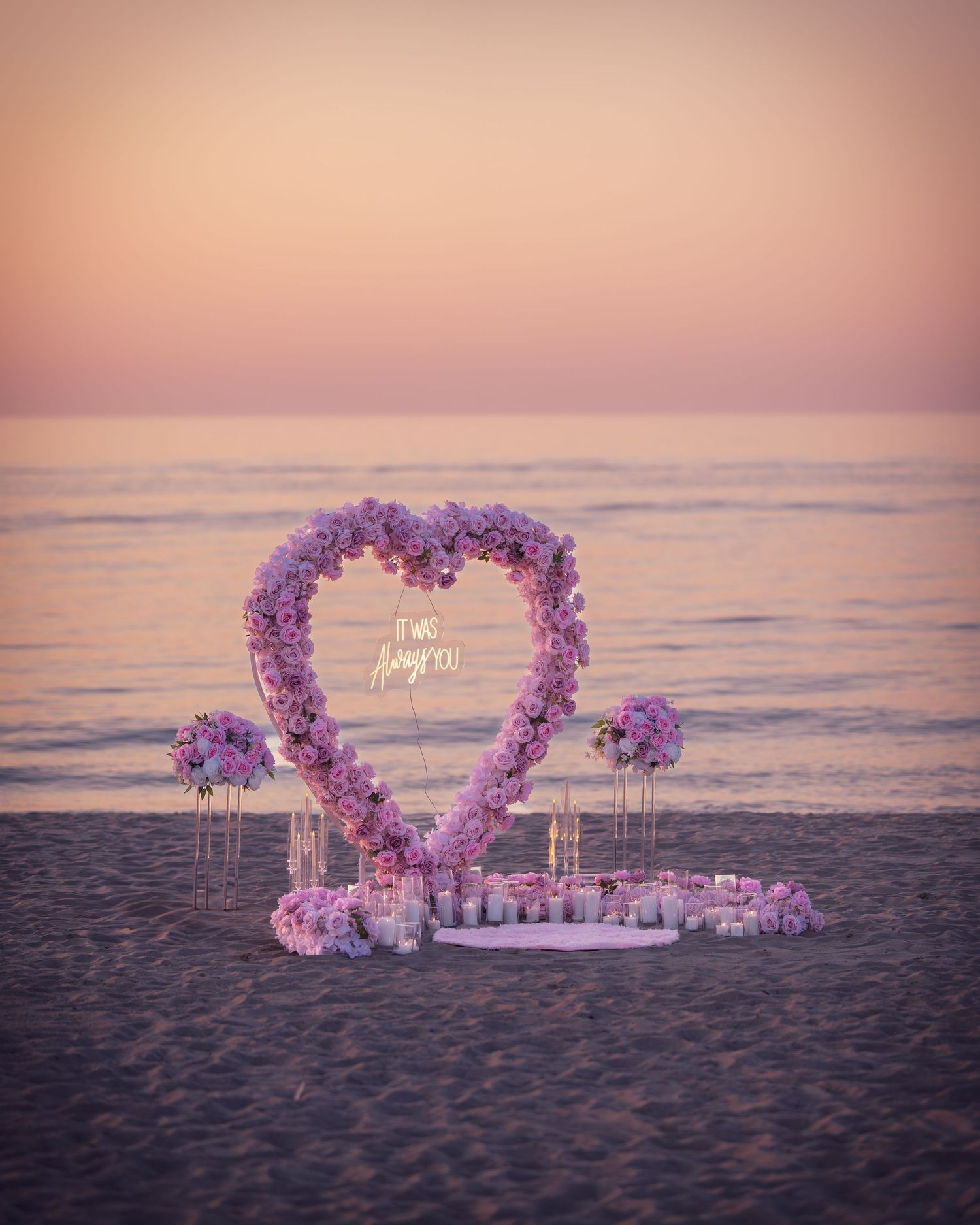 Romantic heart-shaped floral arch with purple flowers and candles set up on a beach at sunset.