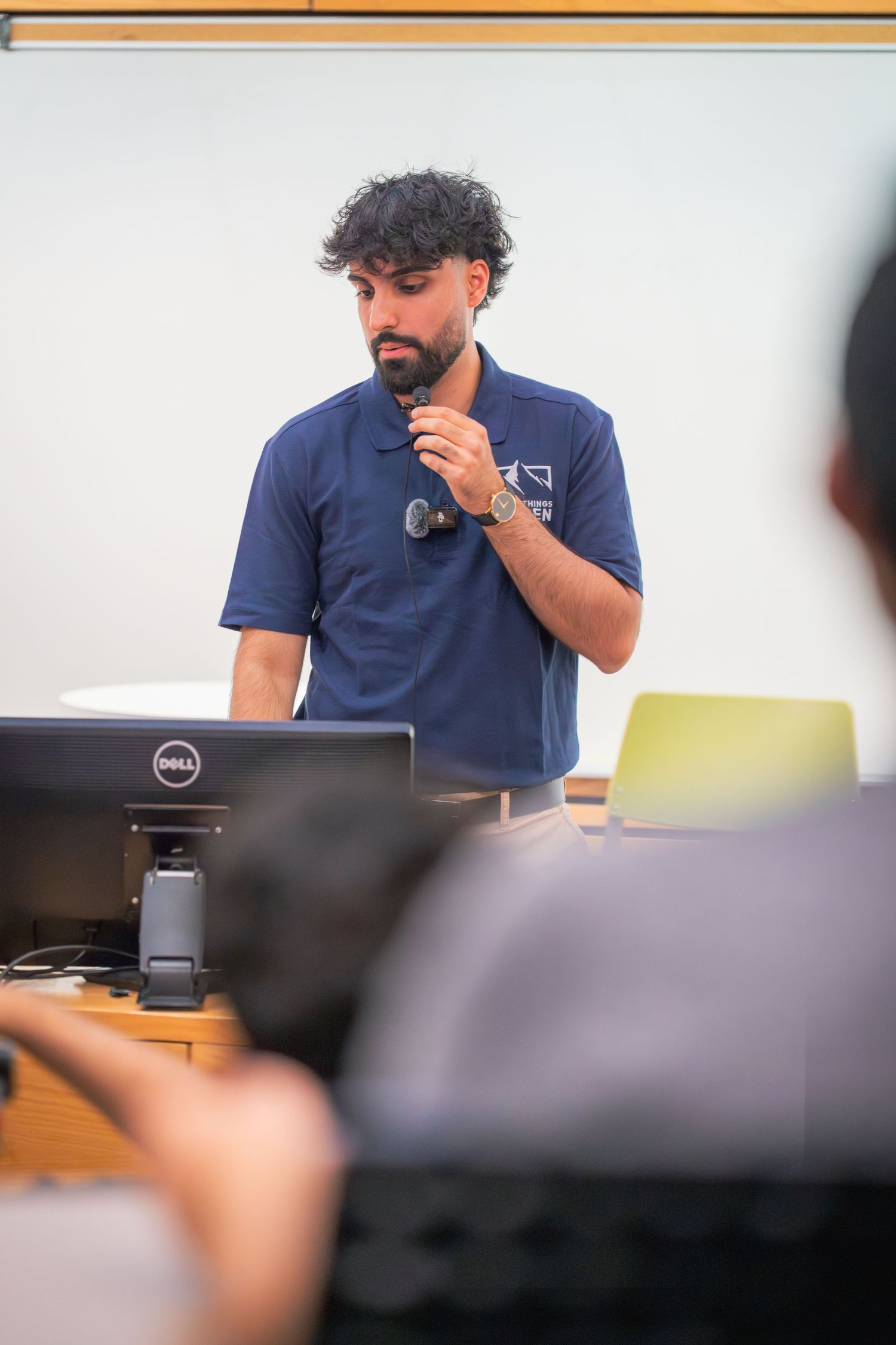 A presenter in a navy blue polo shirt speaks while seated at a desk during a meeting.