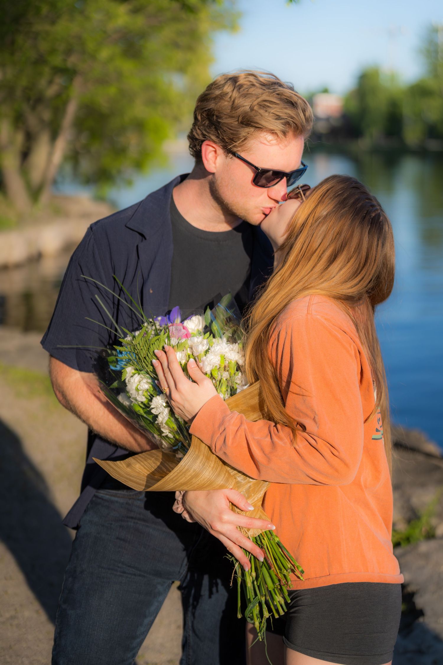 Man kissing woman holding a bouquet of flowers near a scenic riverside at golden hour.