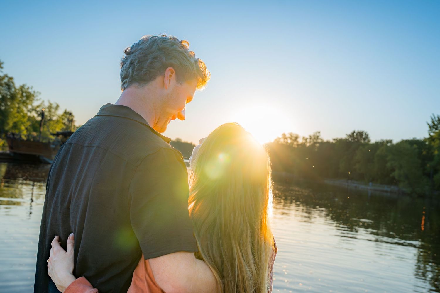 Romantic couple embracing by a lake at golden sunset with warm sun flare glowing between them.