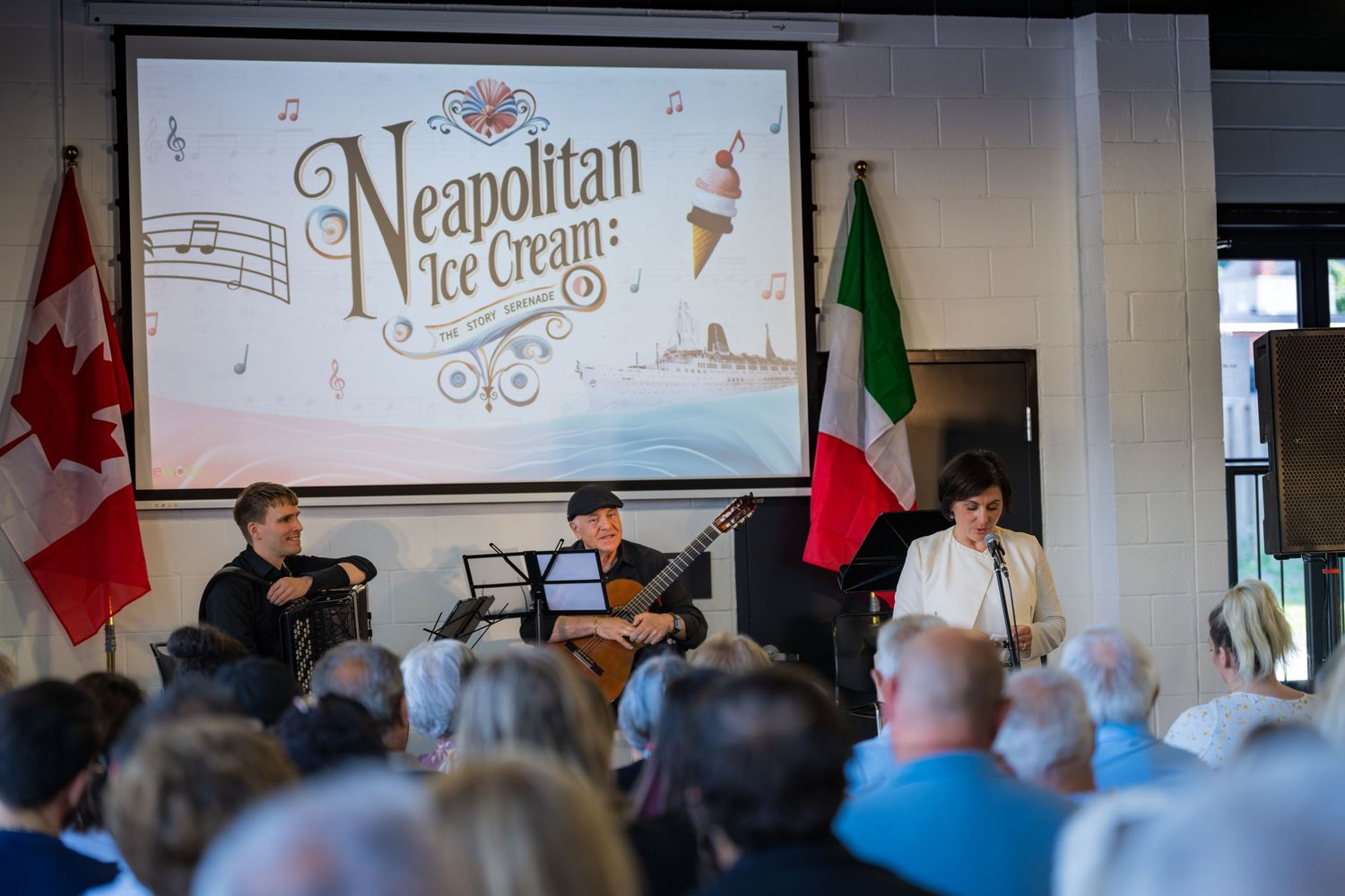 Live musical trio performs at Neapolitan Ice Cream show with audience seated before a decorative backdrop.