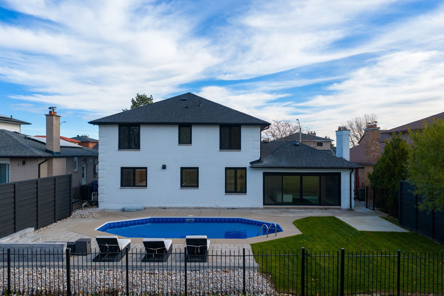 Aerial view of a backyard swimming pool with black metal fencing and white house surrounded by landscaping.