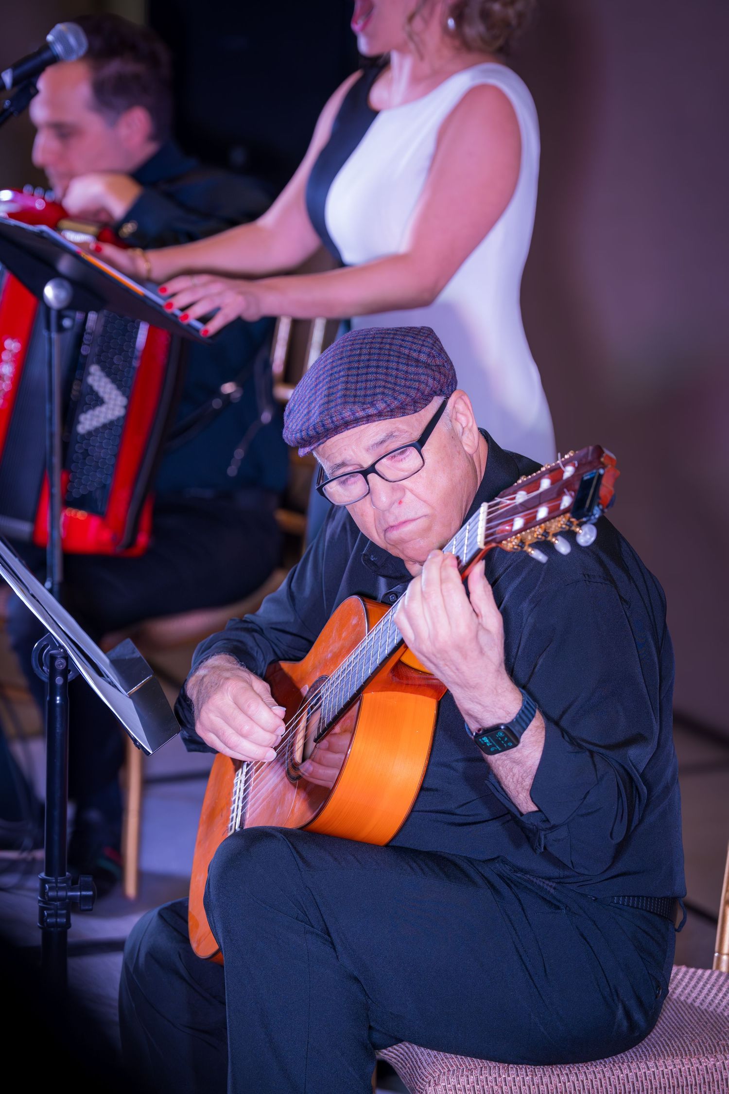 An elderly guitarist in a cap and glasses plays acoustic guitar during a live performance on a dimly lit stage.