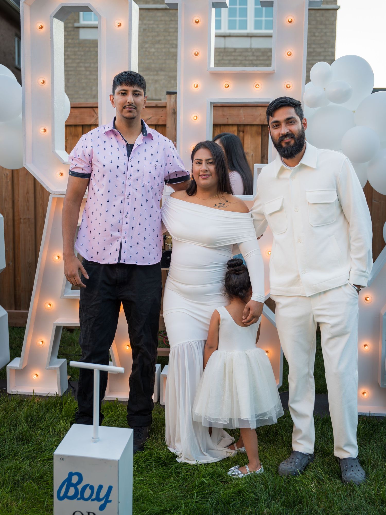 Two men and a pregnant woman in white pose with a toddler at a gender reveal party.