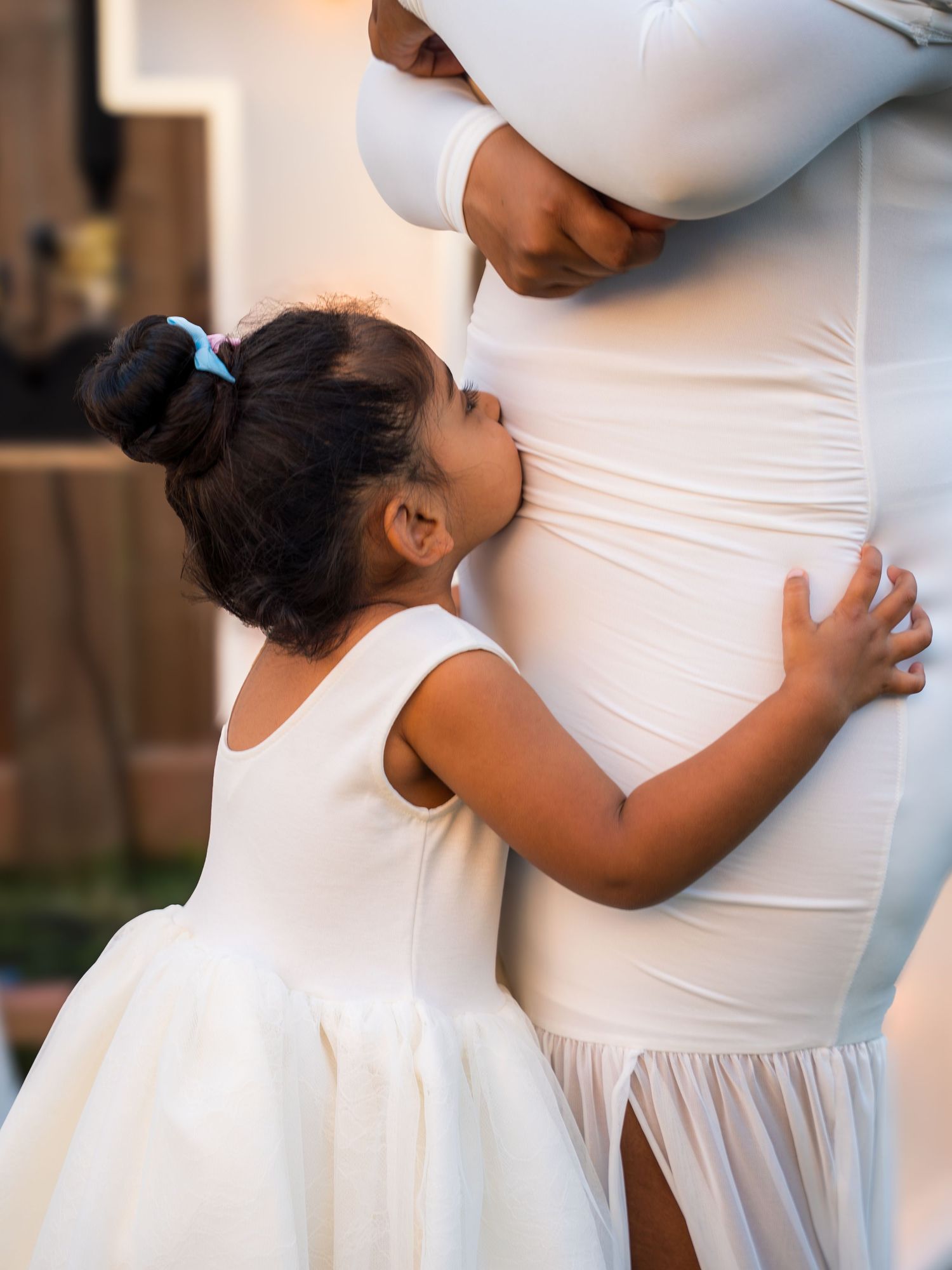 Young girl in white dress kissing pregnant mother's belly during maternity photoshoot.