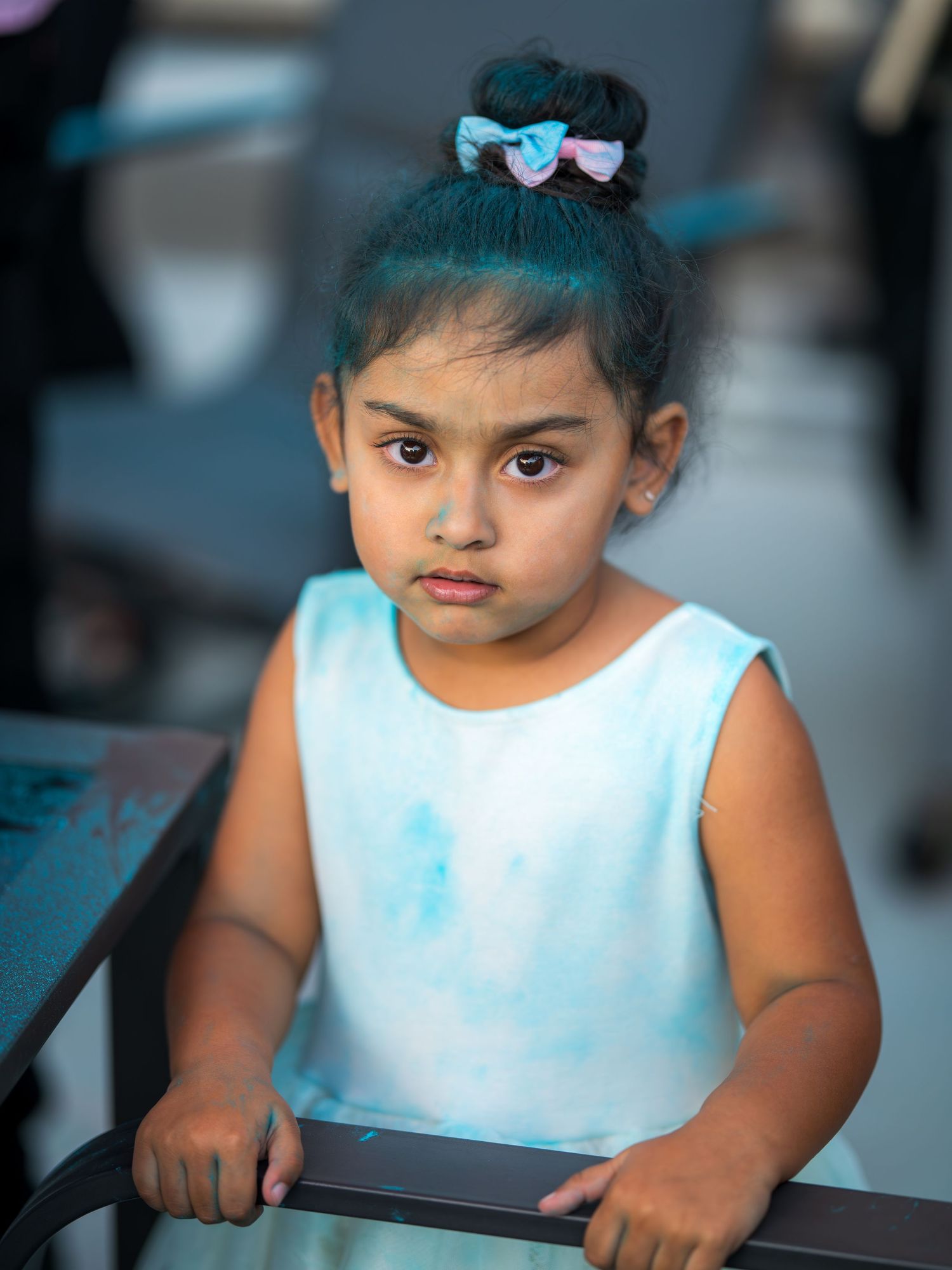 Young girl with teal-tipped hair in blue tie-dye dress looking seriously at camera at outdoor celebration.