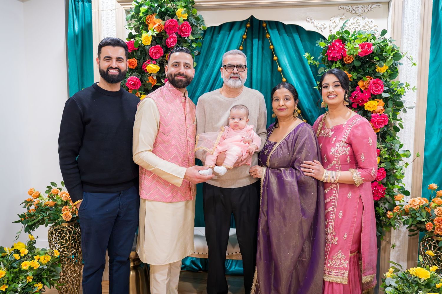Multigenerational family group portrait with newborn baby at vibrant floral and teal draped Indian celebration backdrop.