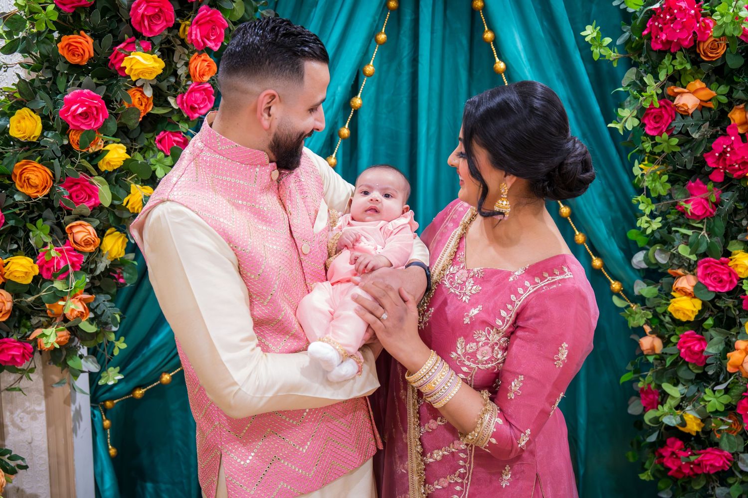 Indian couple in matching pink traditional outfits lovingly gazing at newborn baby at colorful floral backdrop event.