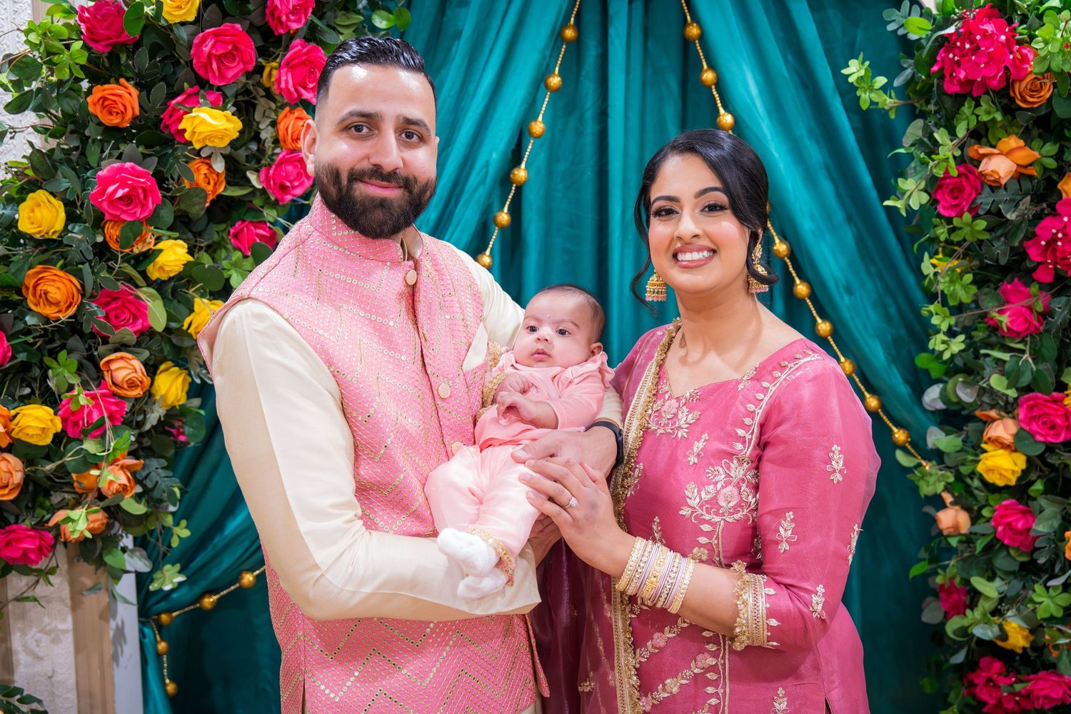 South Asian family in pink traditional attire holding newborn baby, posing before colorful floral backdrop.